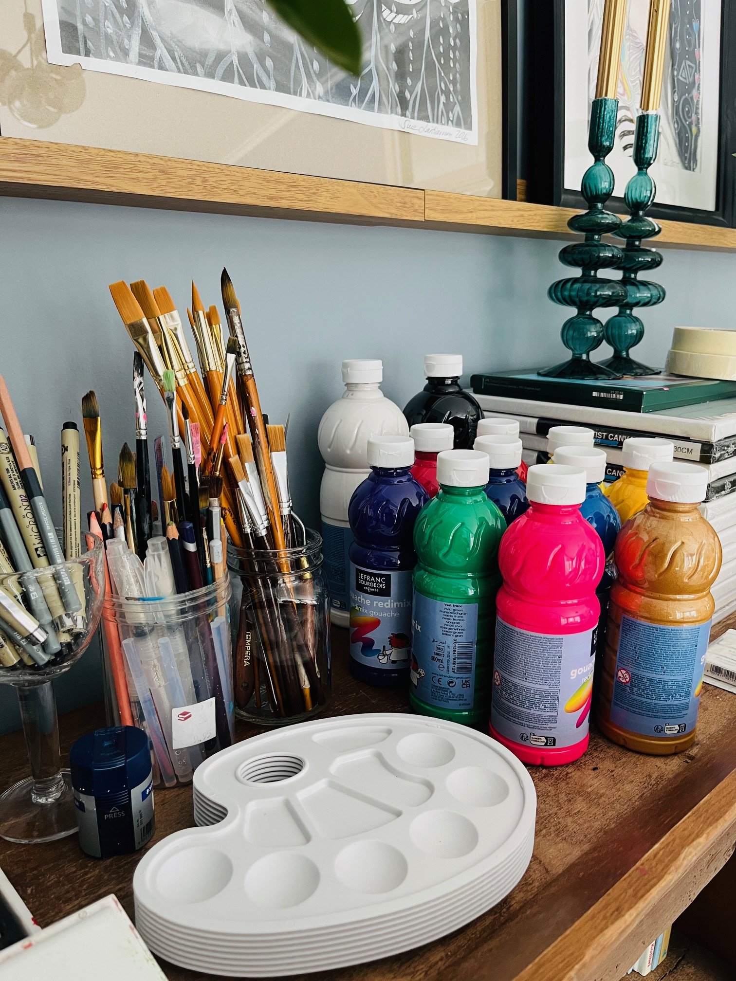 Art supplies including paintbrushes and bottles of gouache paint, a white paint palette, and decorative objects on a wooden desk.
