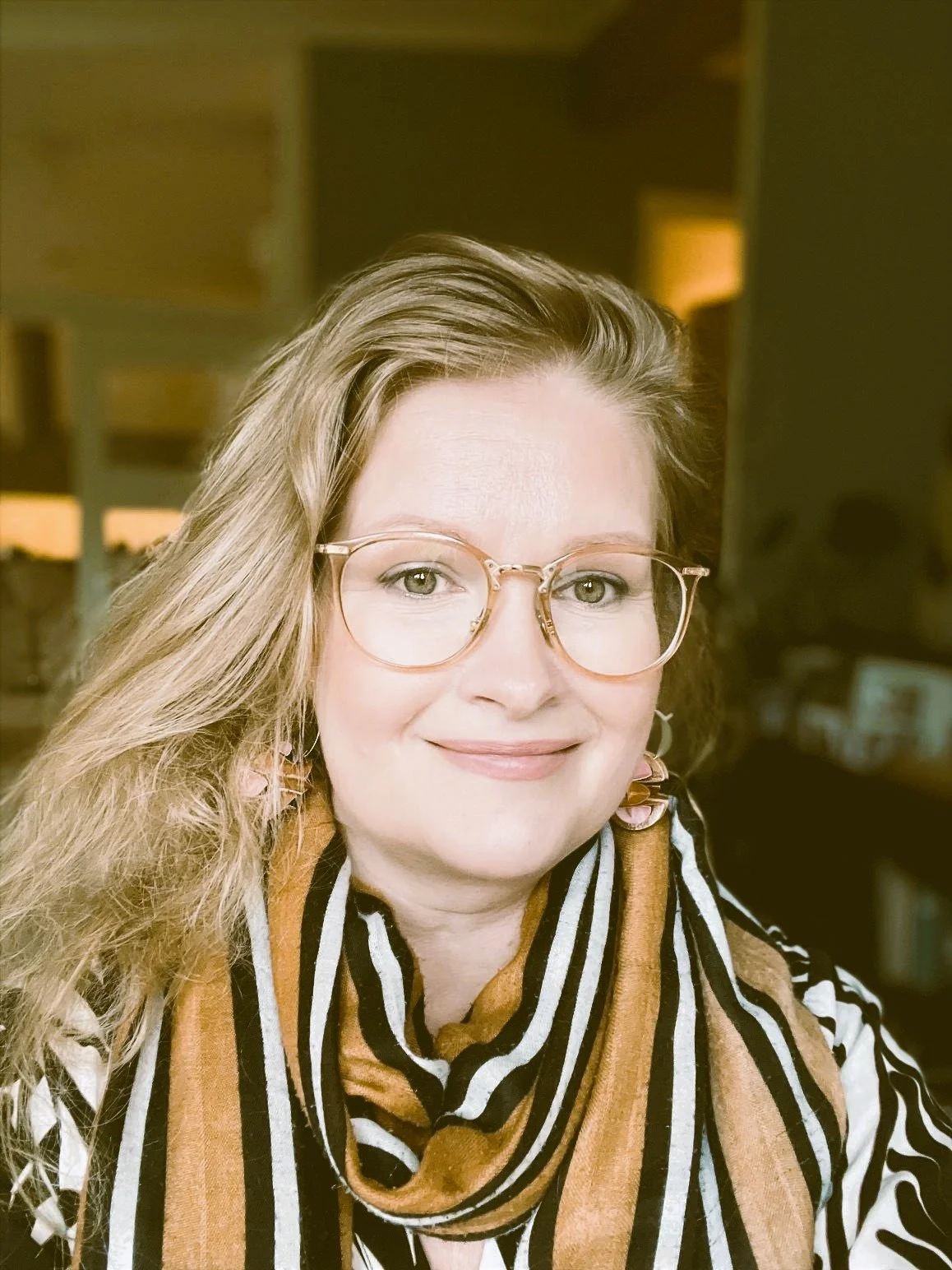 Portrait of a woman with long, wavy hair, wearing glasses, a striped patterned shirt, and a multicolored scarf, smiling in an indoor setting.