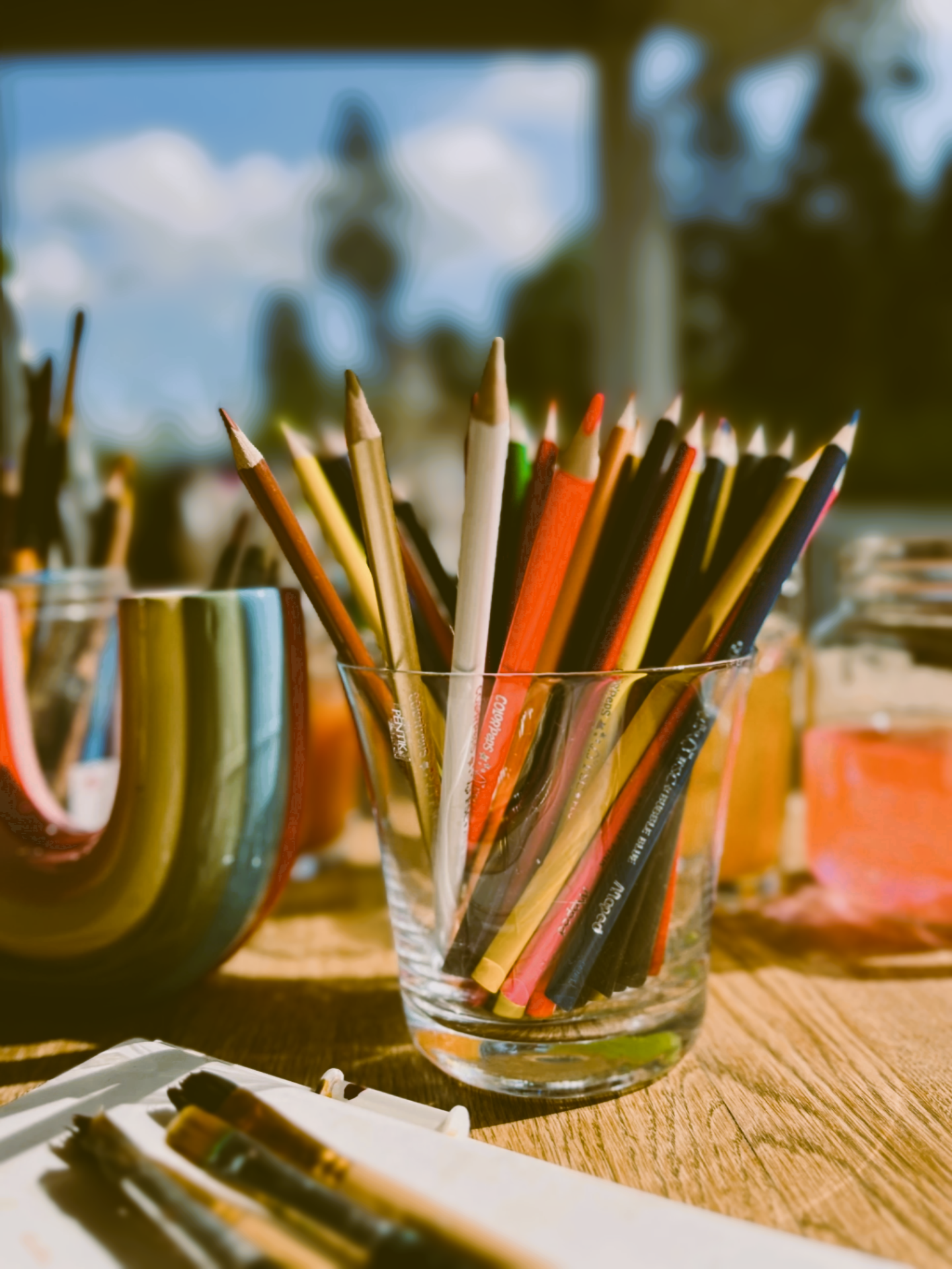 A glass container filled with colored pencils on a wooden table, with additional art supplies and a blurred background of an outdoor scene.