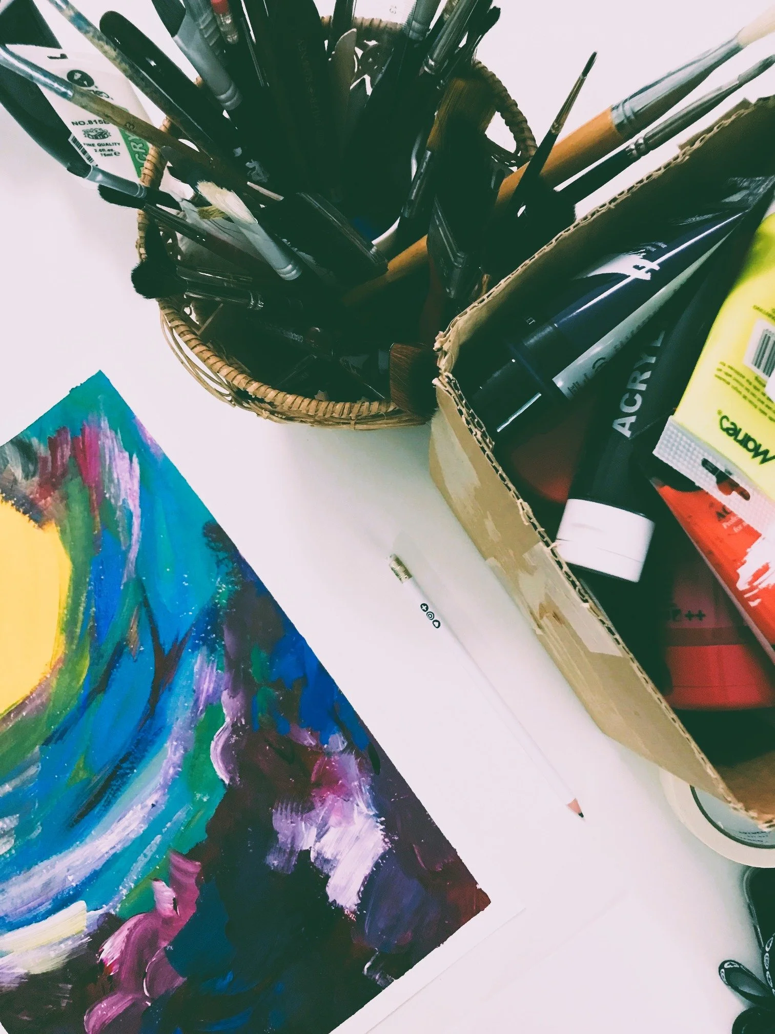 Art supplies on a white table, including a paper with colorful abstract painting, a white pen, and a container holding various brushes and markers.