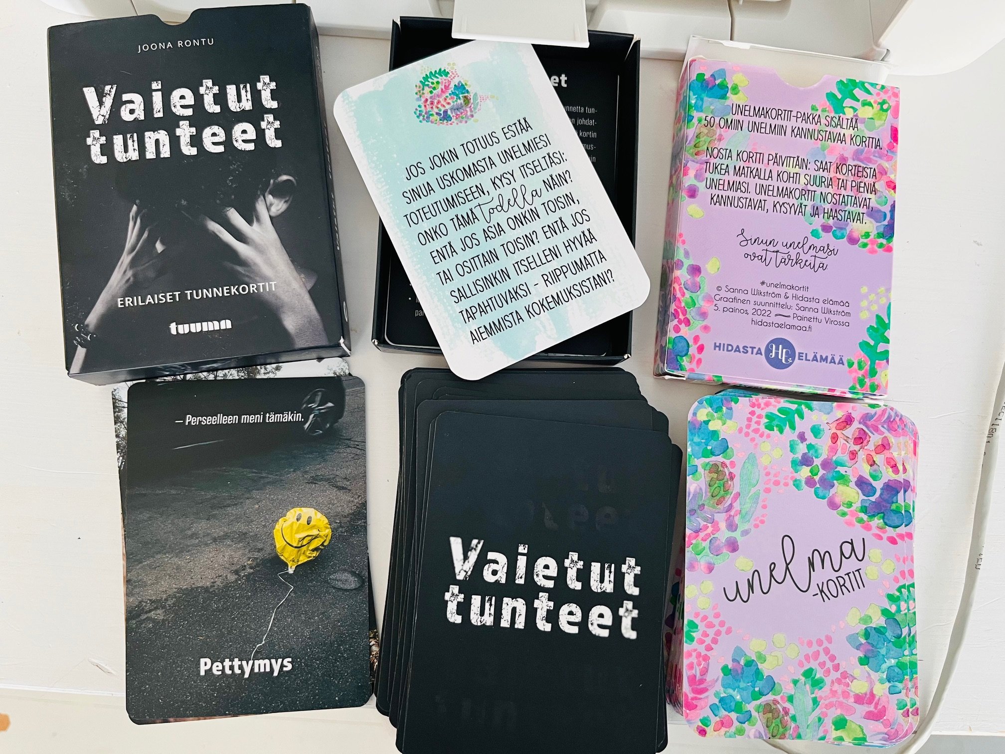 Collection of Finnish self-help cards and booklets, including titles such as "Vaietut tunteet" and "Unelmakortit", with colorful and black-and-white designs placed on a white surface.