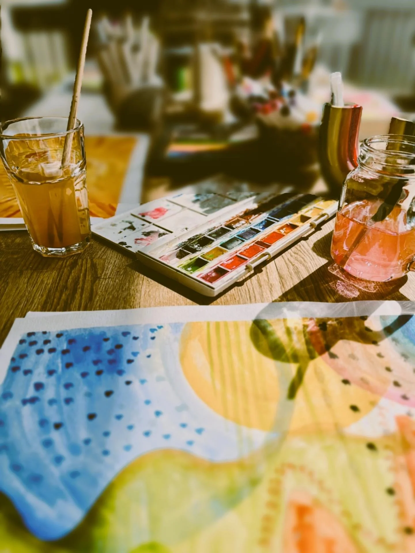 A table with a watercolor palette, a glass of pink beverage, a jar with a pink drink, and colorful artwork in the foreground.