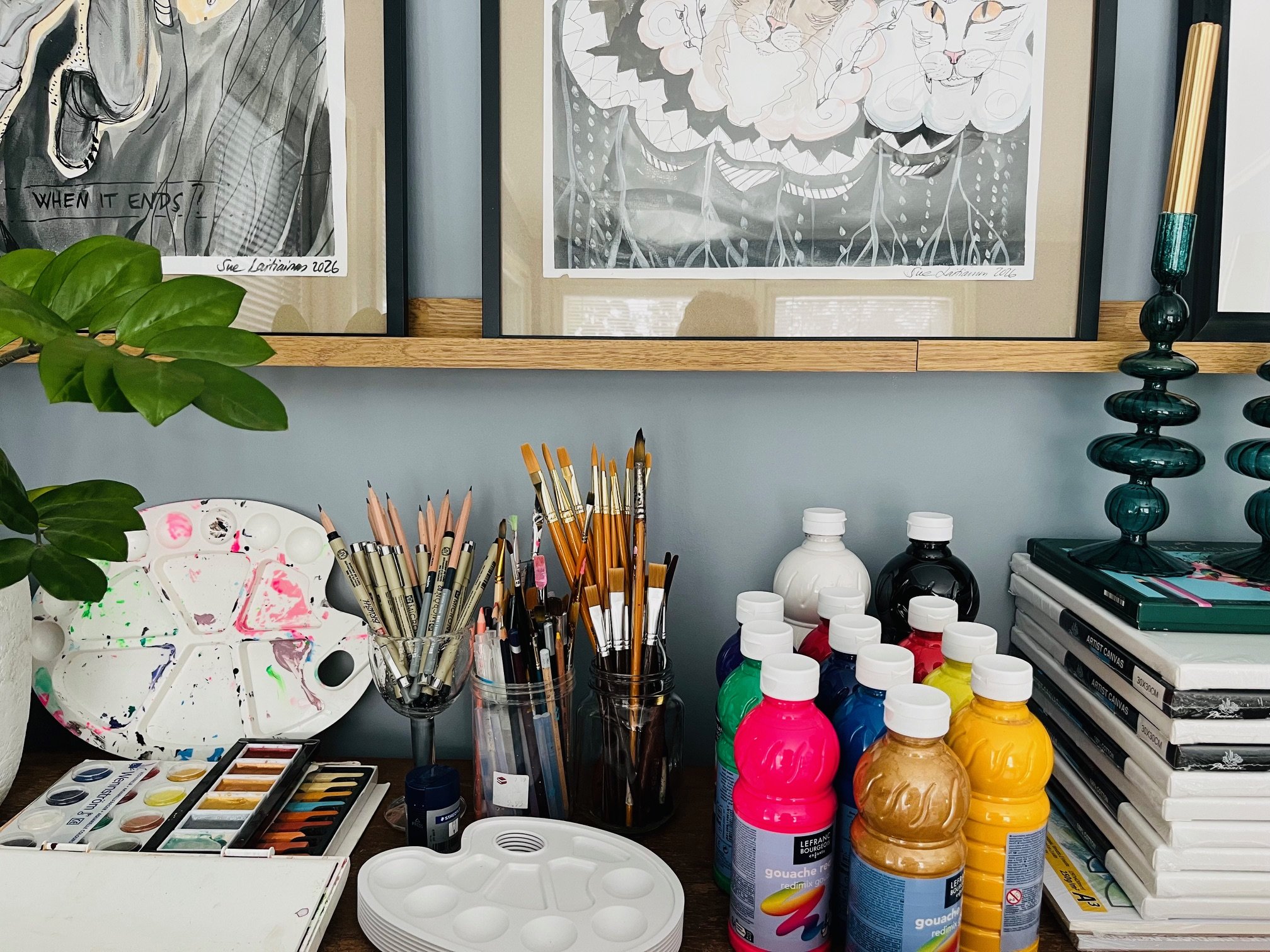 Art supplies and materials on a desk, including paint bottles, brushes, a palette, watercolor sets, sketchbooks, and framed artworks on the wall.
