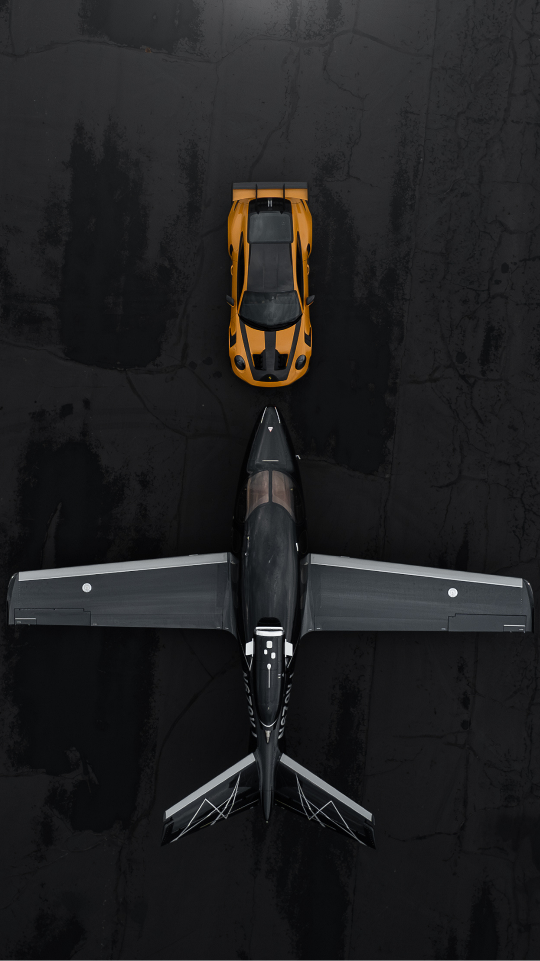 An aerial view of a yellow sports car parked behind a black fighter jet on a dark runway.