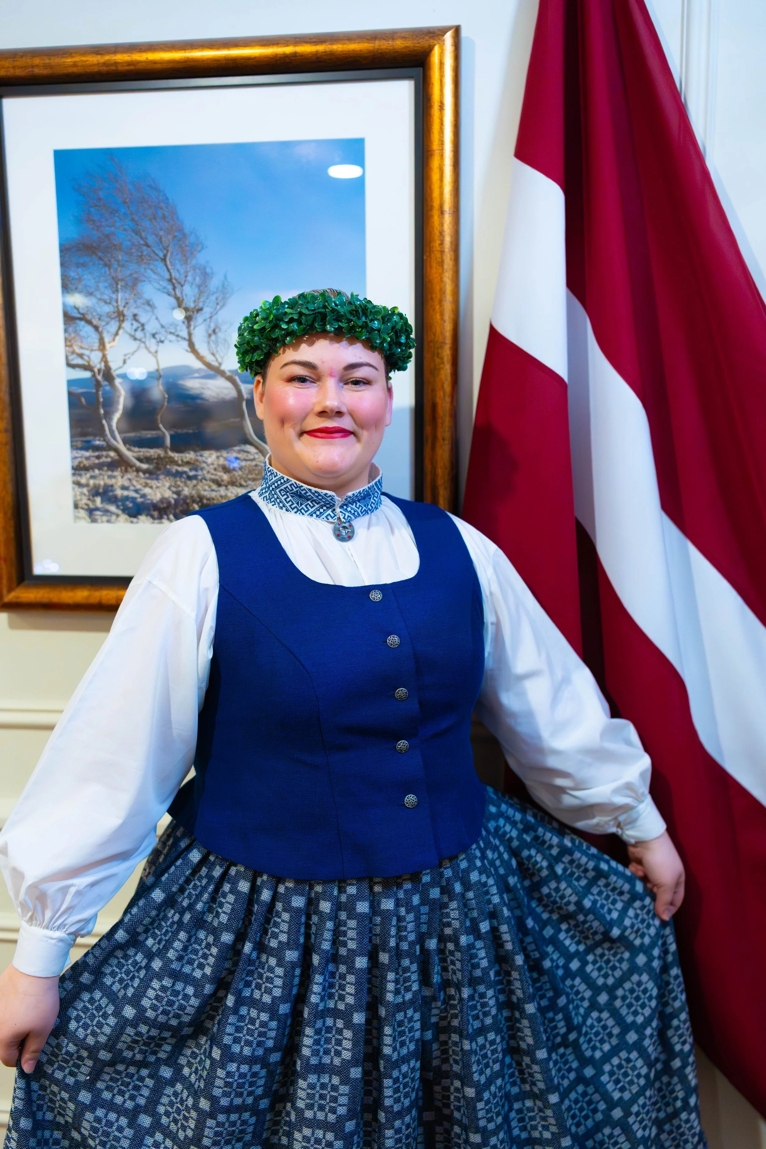 A woman in traditional Norwegian attire standing near a Norwegian flag and a framed landscape picture.