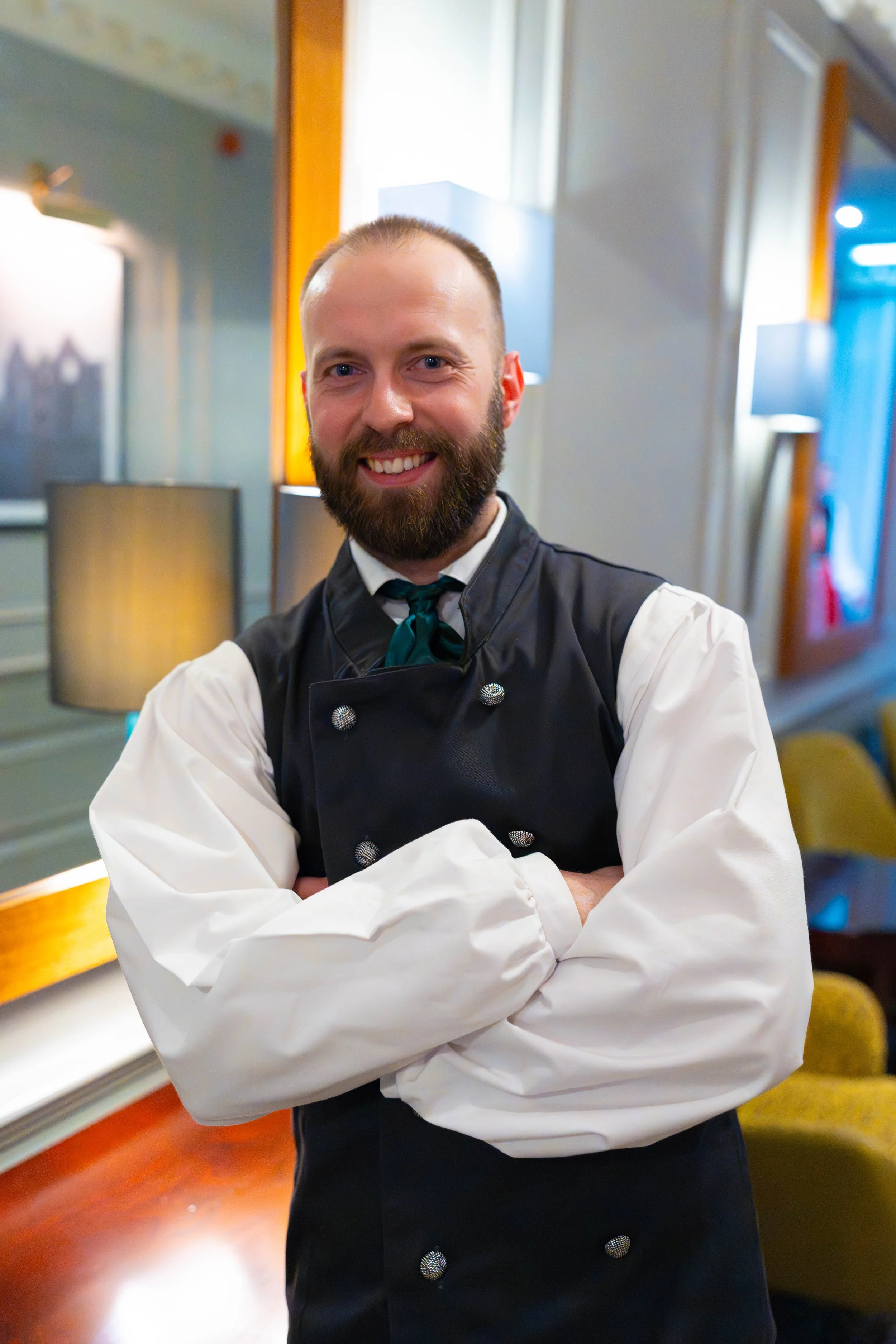 A smiling man with a beard, wearing a white chef's coat and black vest, standing with arms crossed in a warmly lit room.