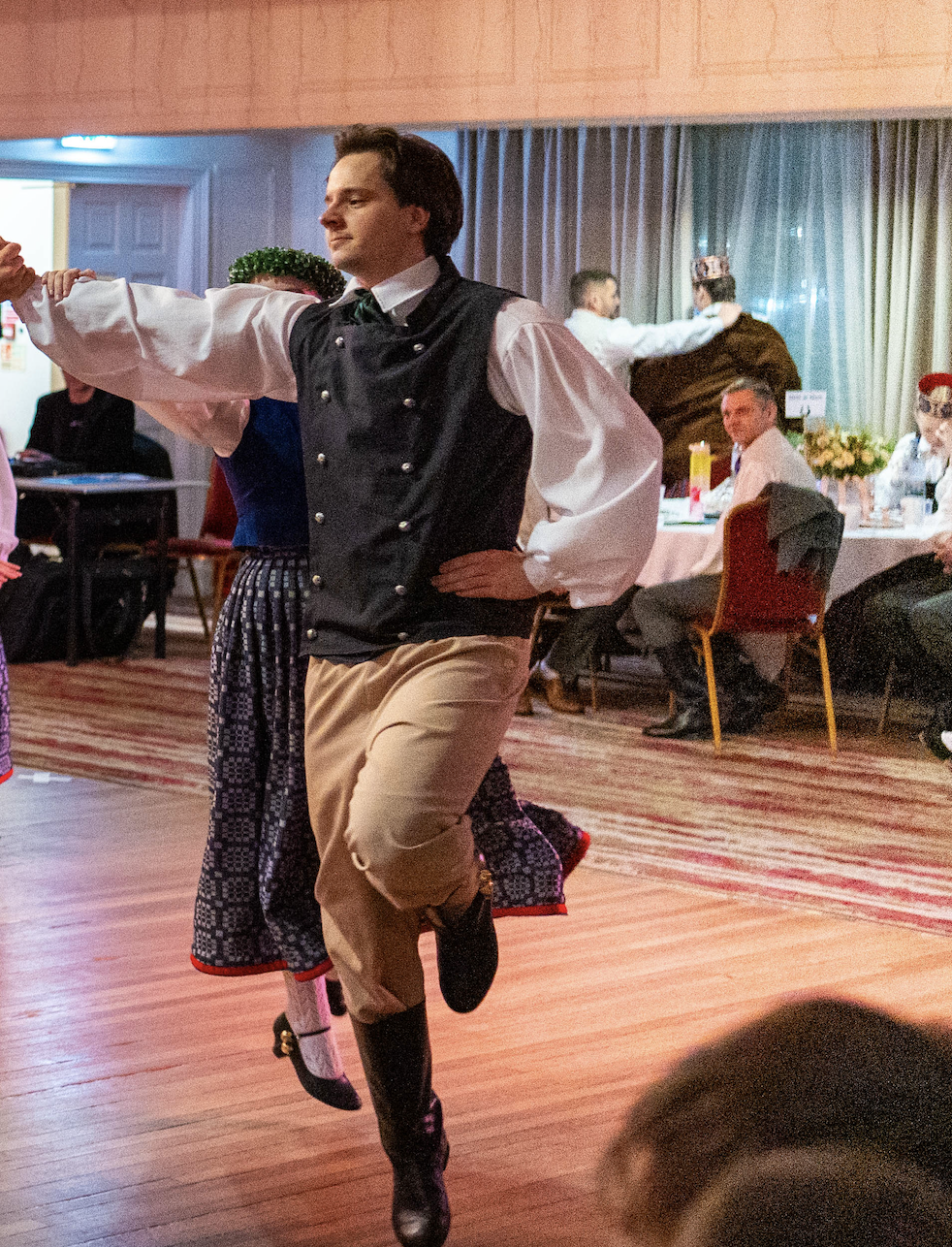 A man wearing traditional Scottish attire, including a black vest and tan trousers, is dancing with his arms outstretched and one leg raised in a ballroom or banquet hall setting.