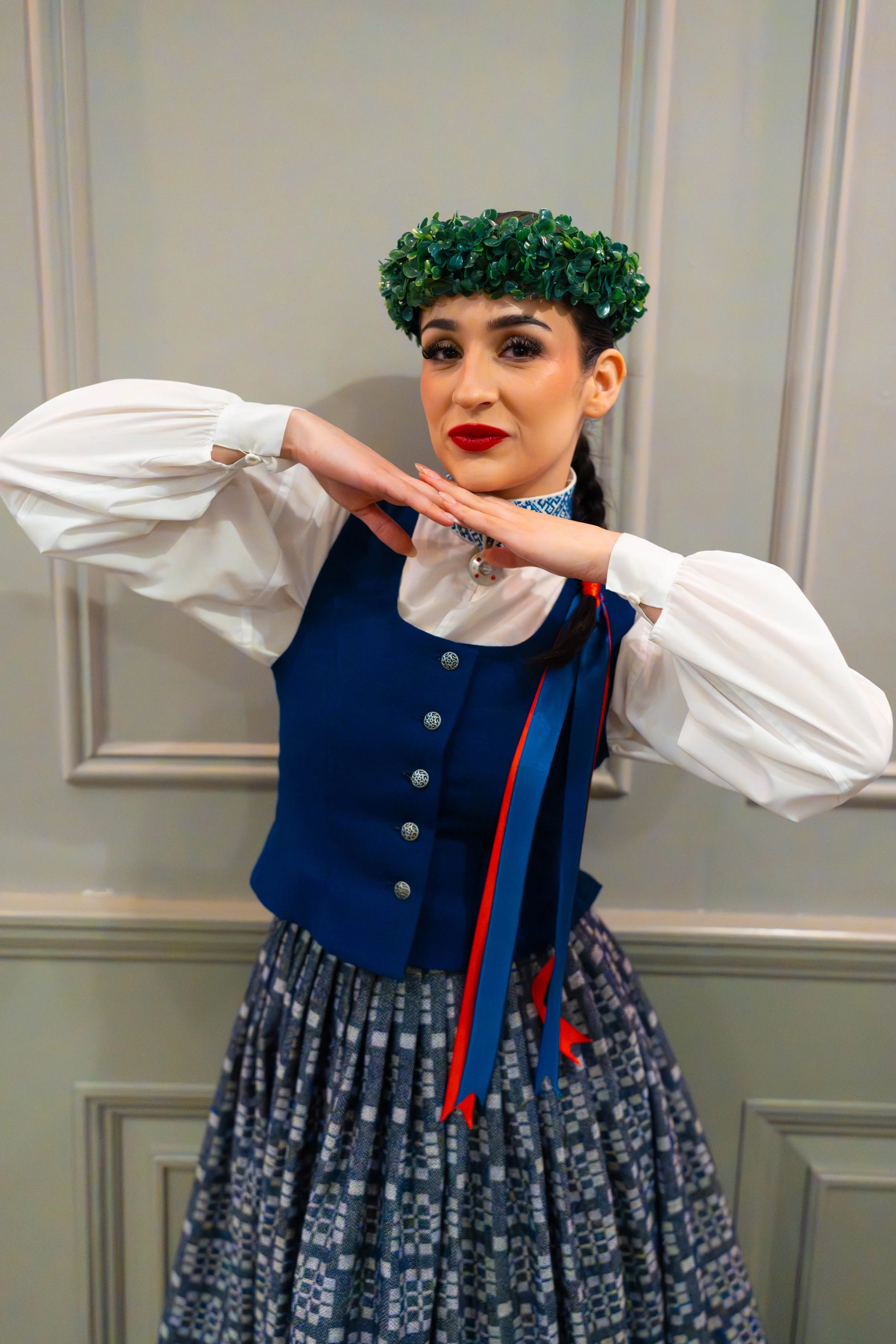 A woman wearing a traditional outfit with a green leafy hat, giving a pose with her hands under her chin, standing in front of a paneled wall.