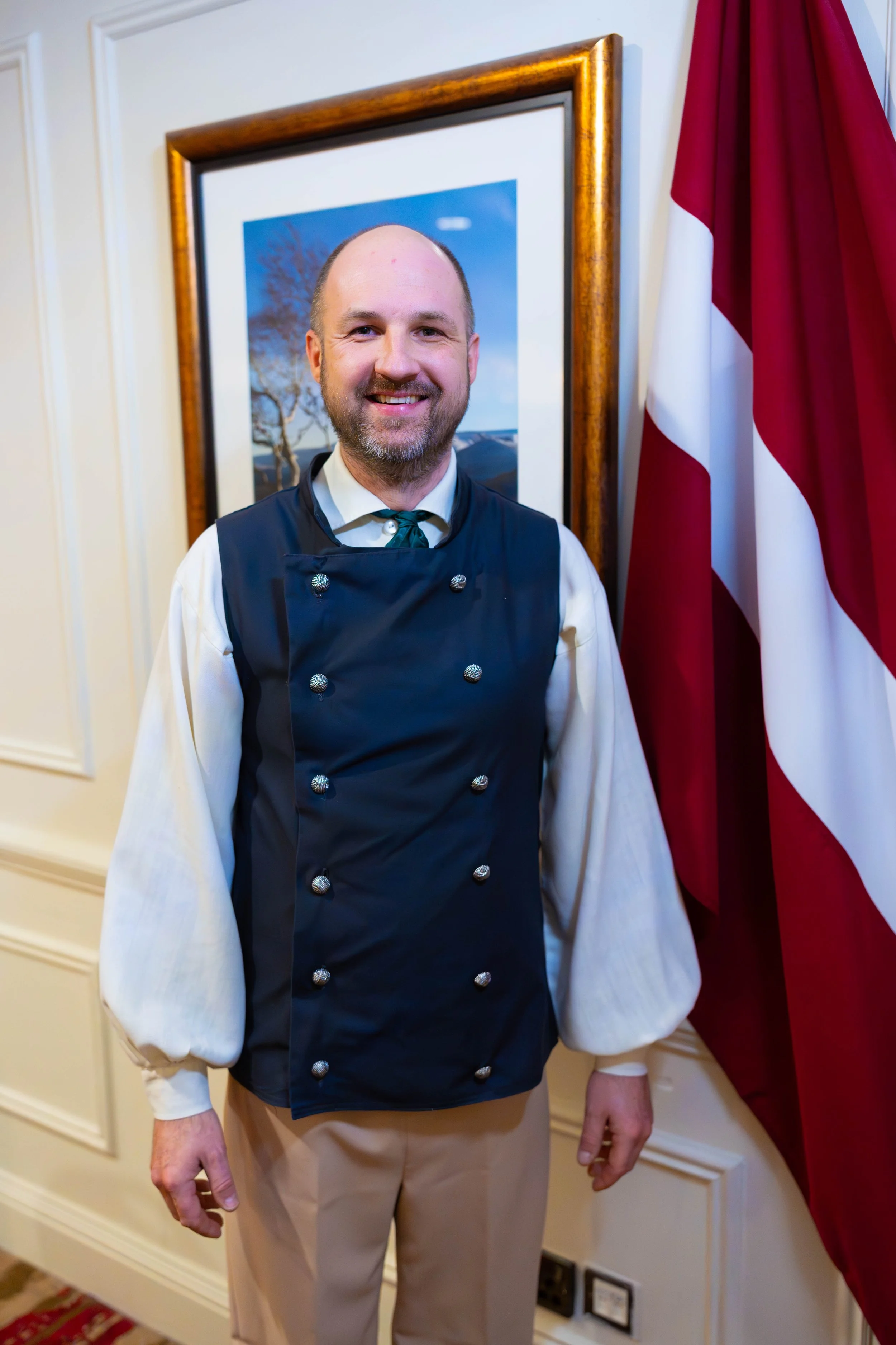 A man in a chef's uniform standing indoors, smiling, with a framed picture of a tree and sky behind him, and a red and white flag on the right side.