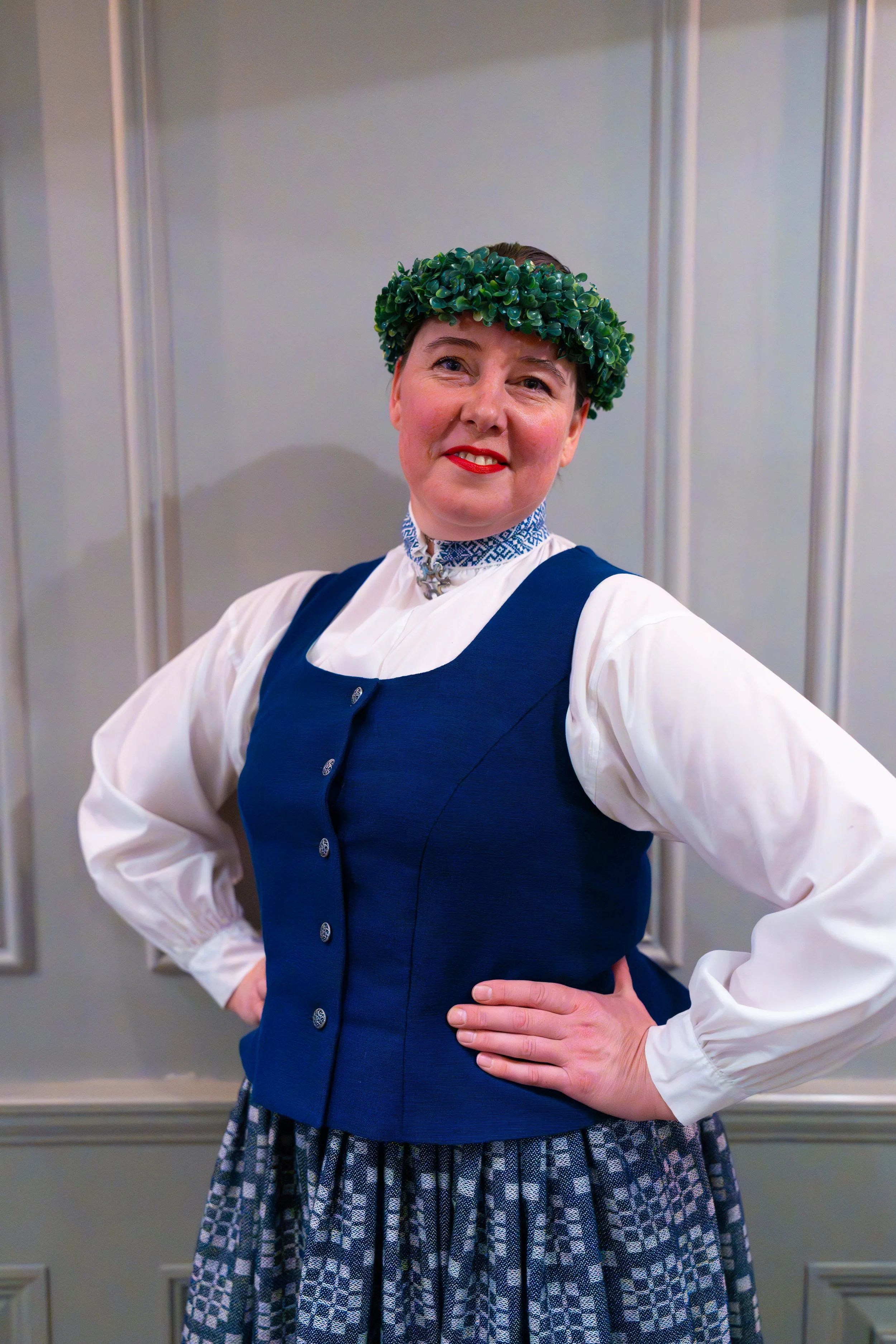 A woman dressed in traditional clothing, wearing a leafy green crown, standing with one hand on her hip in front of a neutral background.