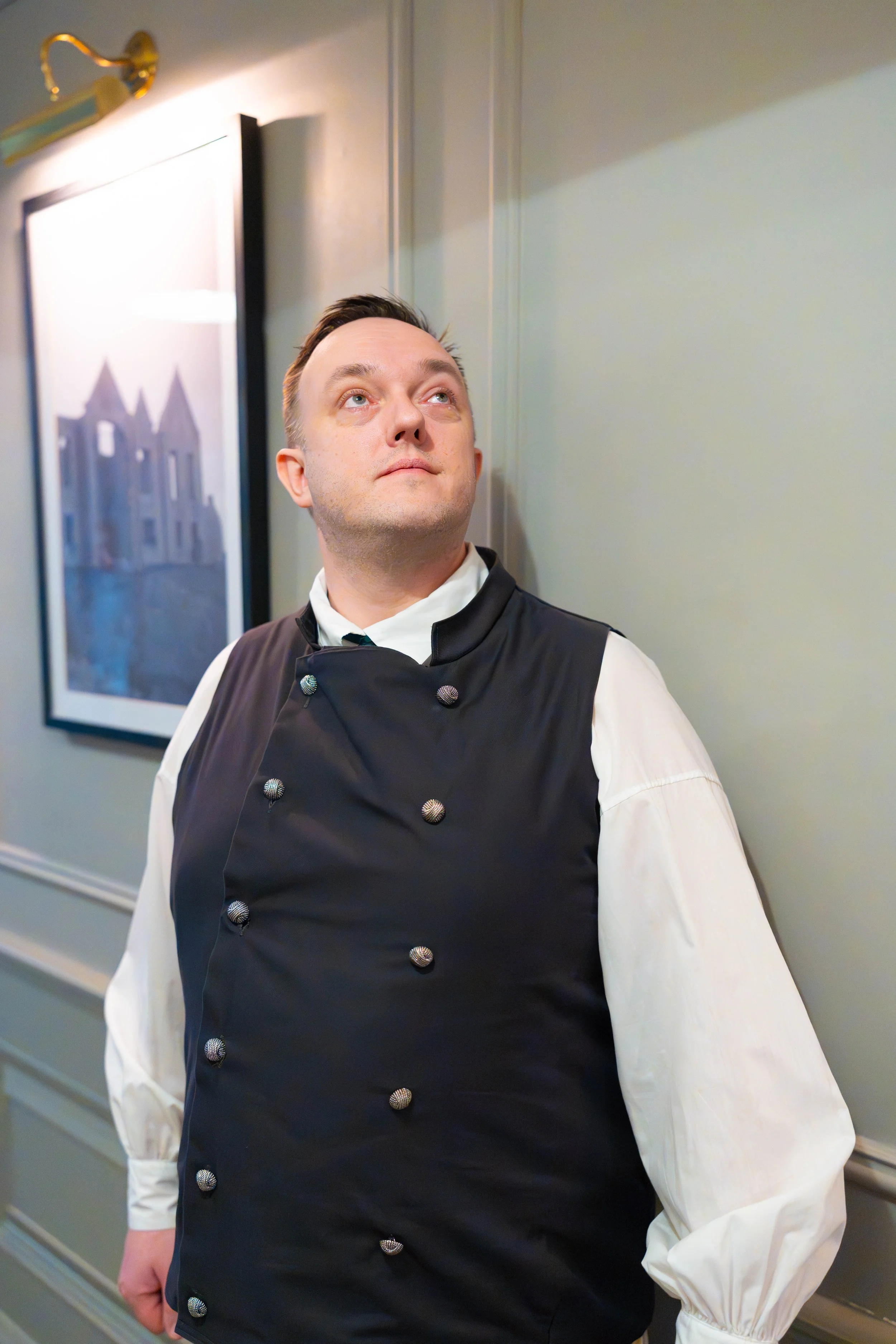 A man dressed as a chef standing in an indoor setting, looking to the side. He is wearing a black double-breasted chef jacket with metal buttons and a white shirt underneath. There is a framed picture on the wall behind him.