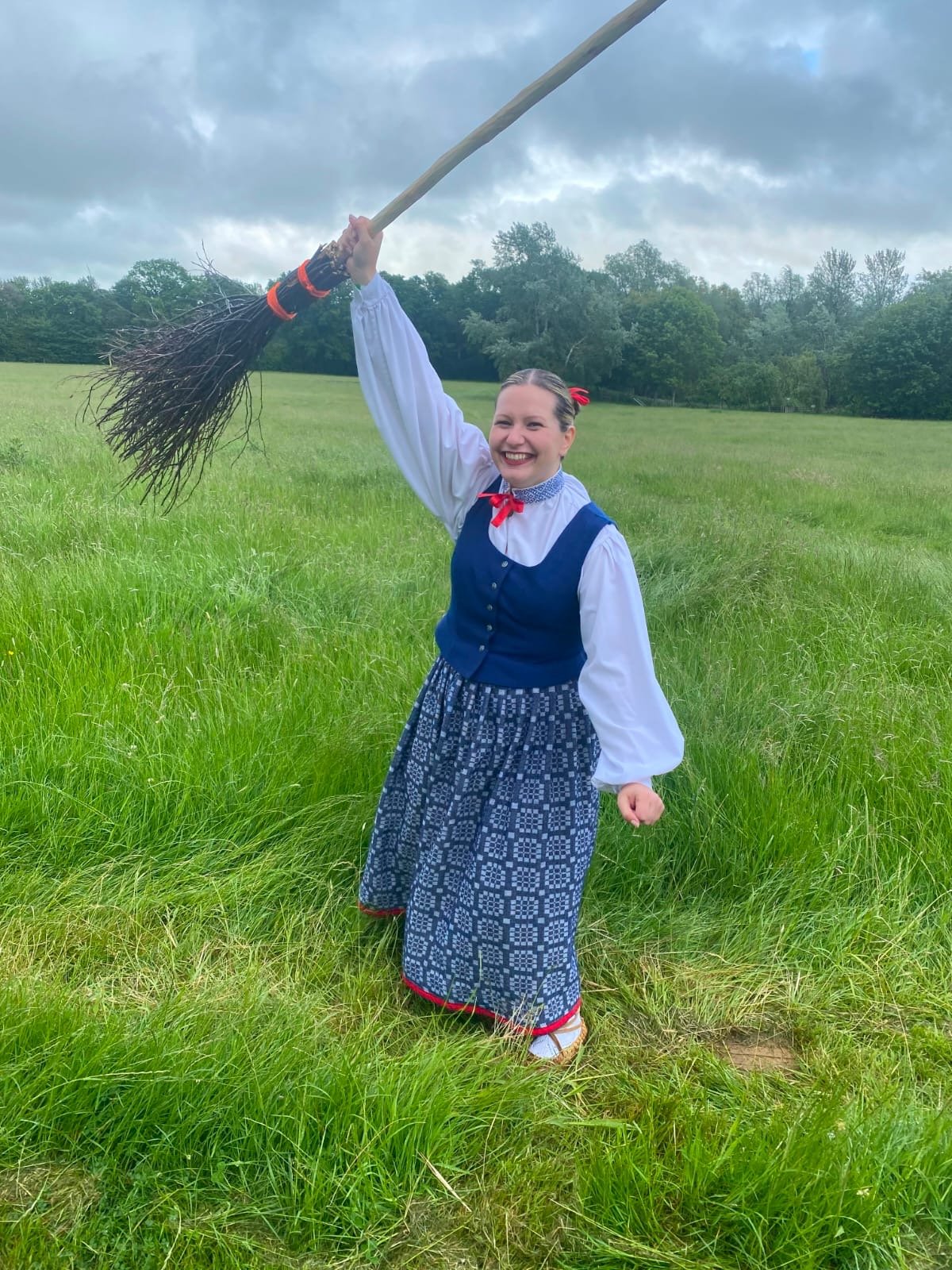 A young woman in traditional clothing holding a broomstick above her head in a green, grassy field under a cloudy sky, smiling joyfully.