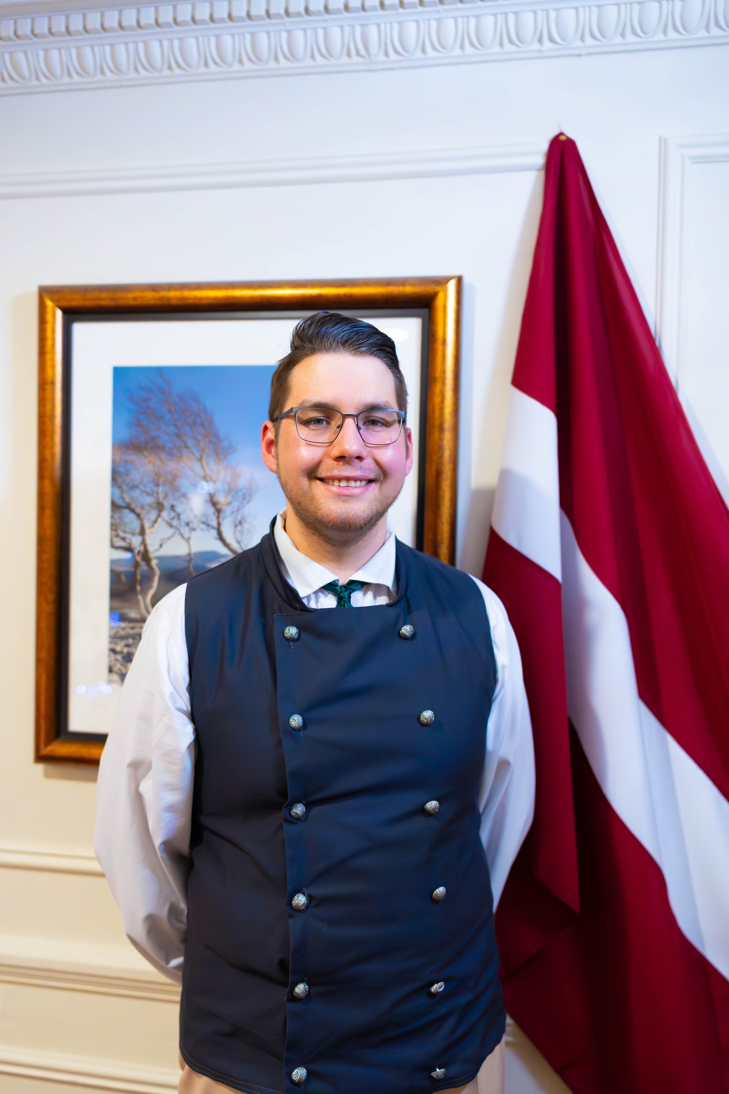 A man dressed as a chef standing in front of a framed picture of trees and a flag with red, white, and blue colors, inside a room with ornate ceiling molding.