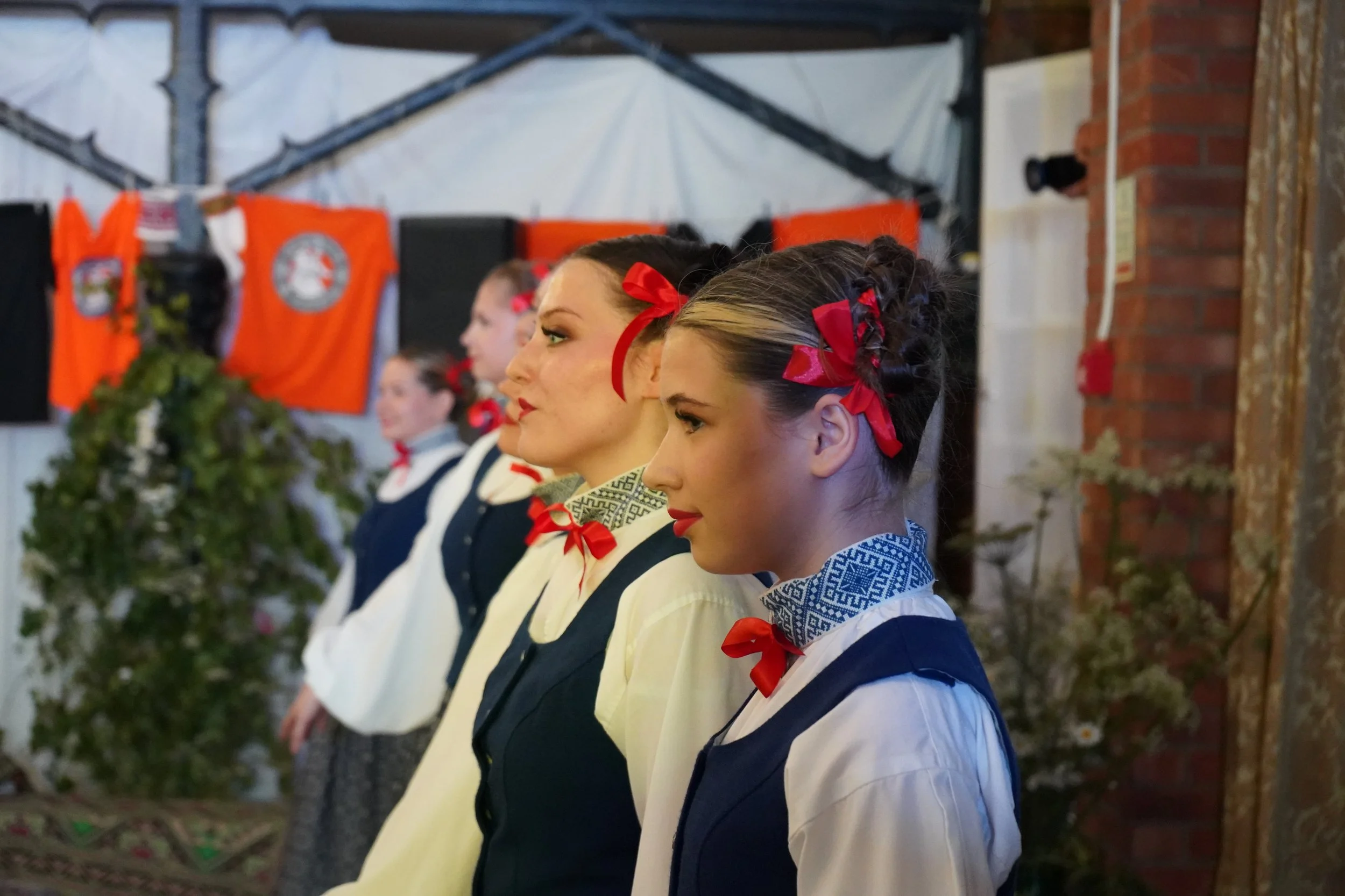 A line of women dressed in traditional Ukrainian attire, standing in profile with their heads slightly bowed, adorned with red ribbons in their hair, inside a room decorated for a cultural event with Ukrainian flags and plants.