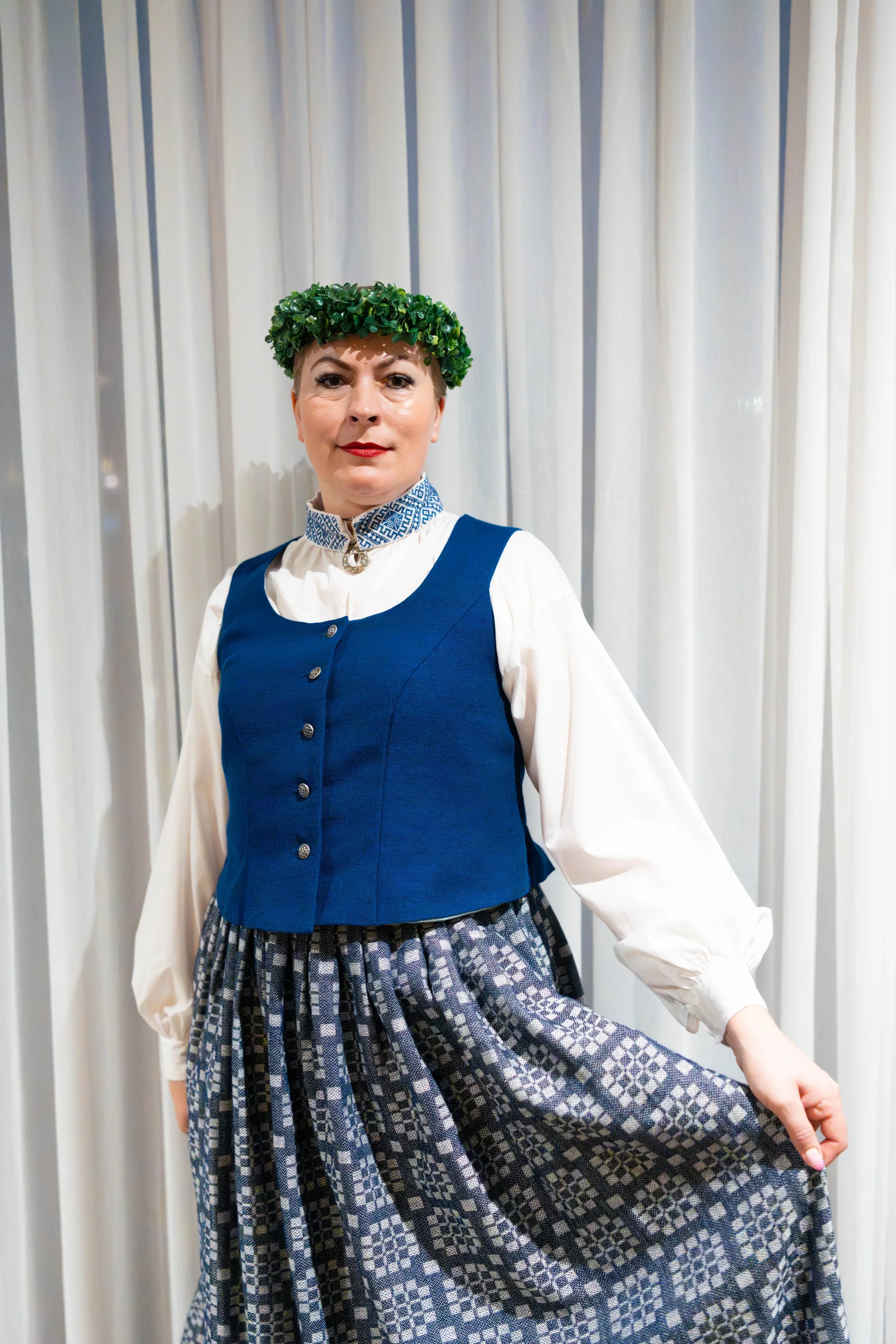 A woman in traditional attire with a green leafy headpiece, white blouse, blue vest, and a patterned skirt, standing in front of white curtains.