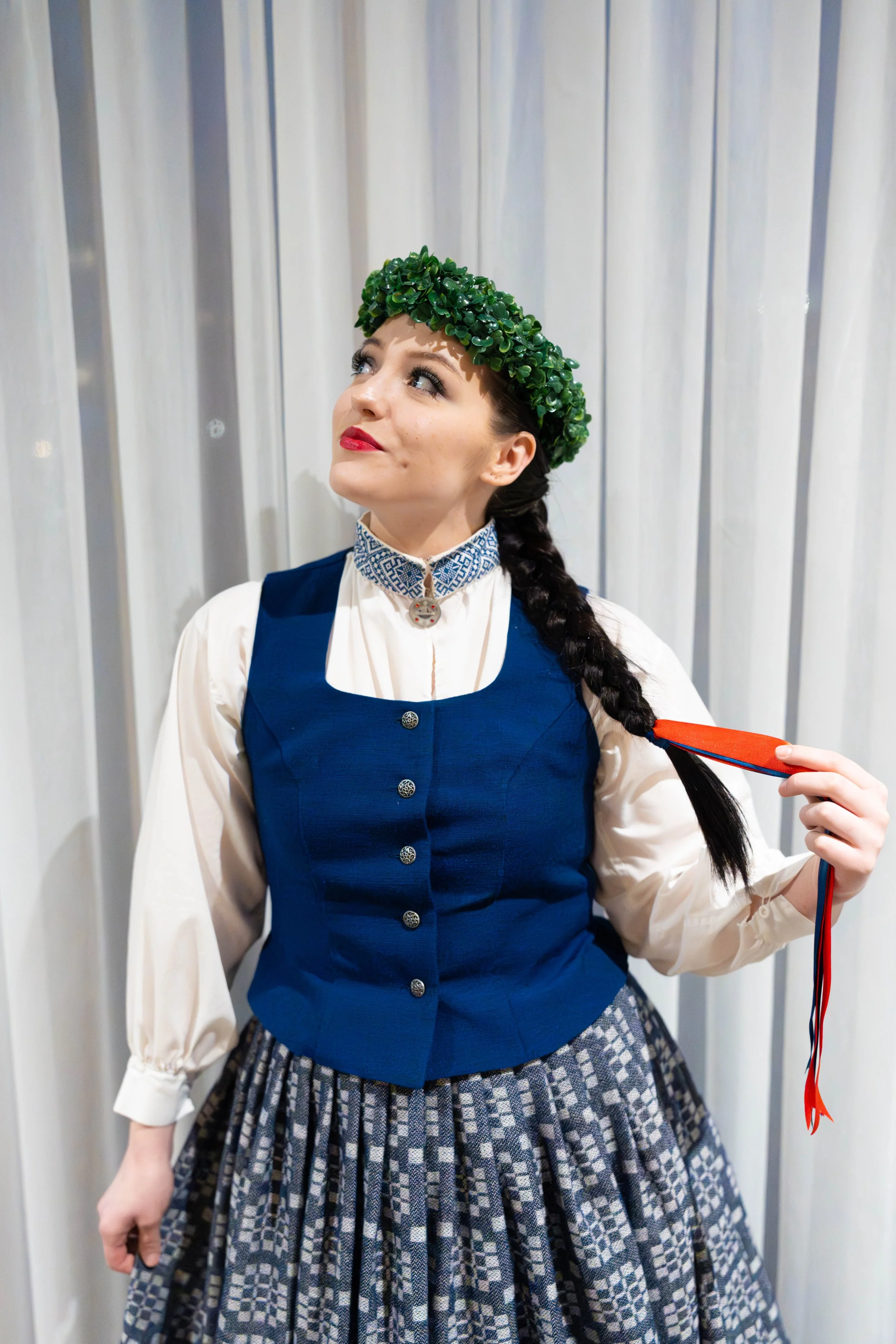 A woman dressed in traditional Latvian attire, wearing a blue vest over a white blouse, a patterned skirt, and a green leafy crown, holding a red and blue ribbon.