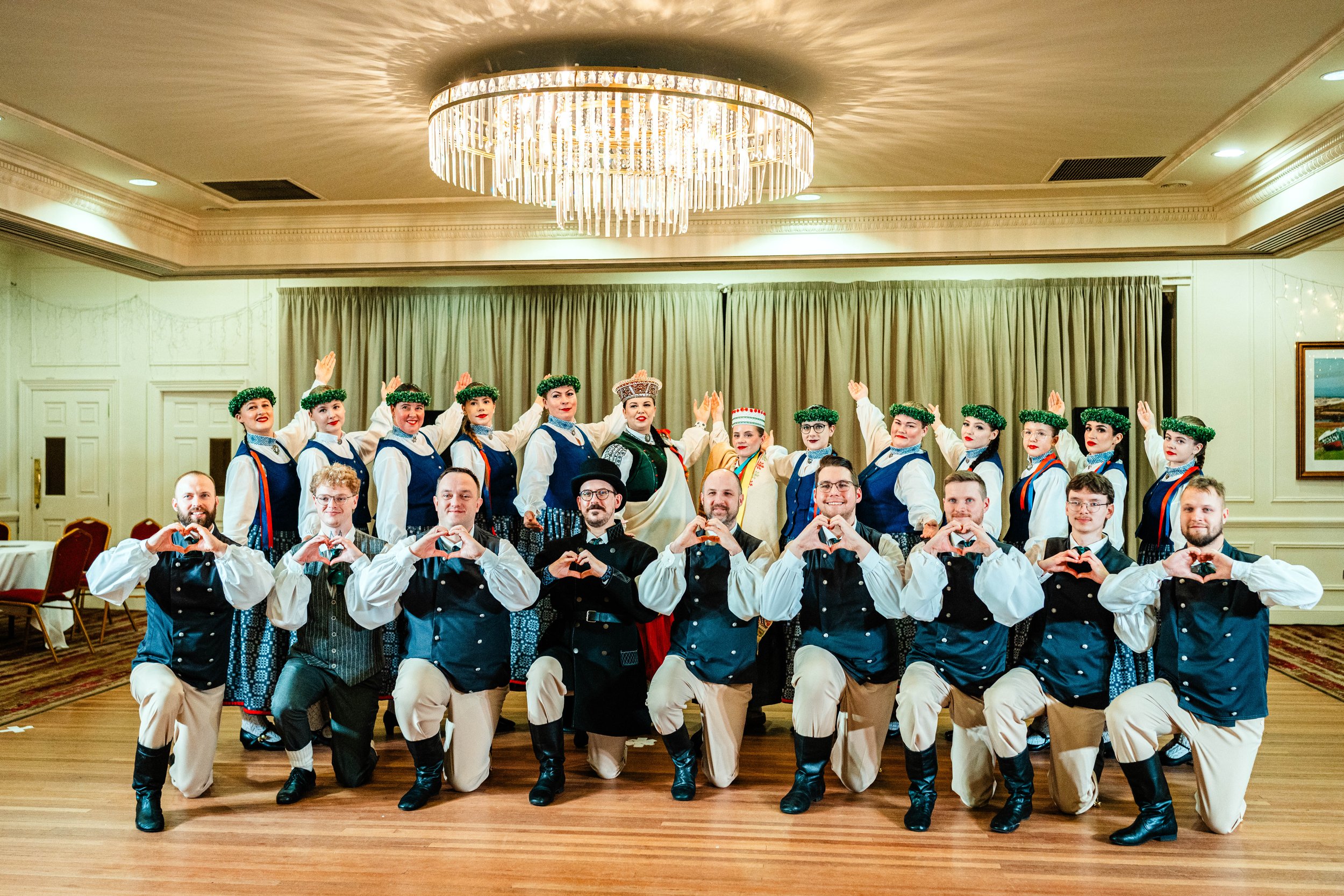 Group of performers in traditional folk costumes posing in a banquet hall, some making heart shapes with their hands, with a chandelier overhead.