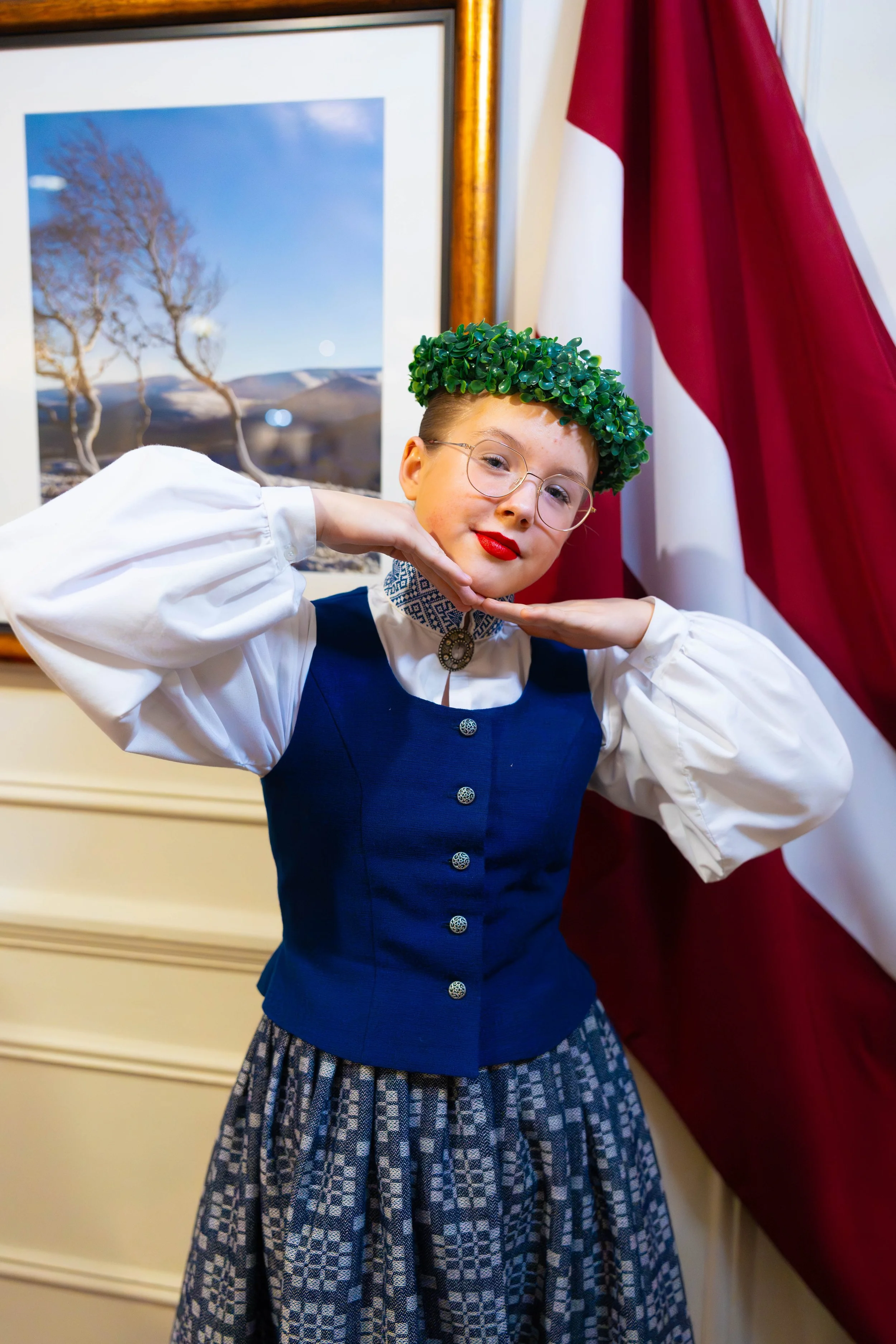 Child wearing a traditional dress with glasses, red lipstick, and a green leafy crown posing with hands on chin in front of a framed landscape picture and American flag.