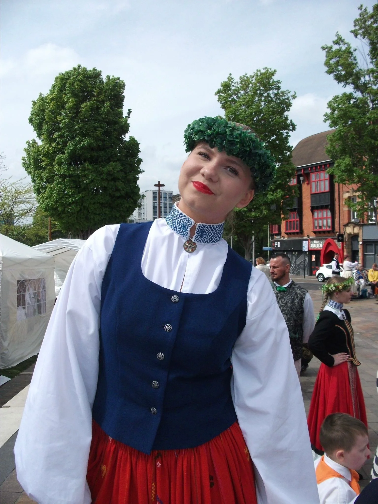 A woman wearing traditional clothing, including a green leafy headpiece and a blue vest over a white blouse, smiling at an outdoor cultural event with people and trees in the background.
