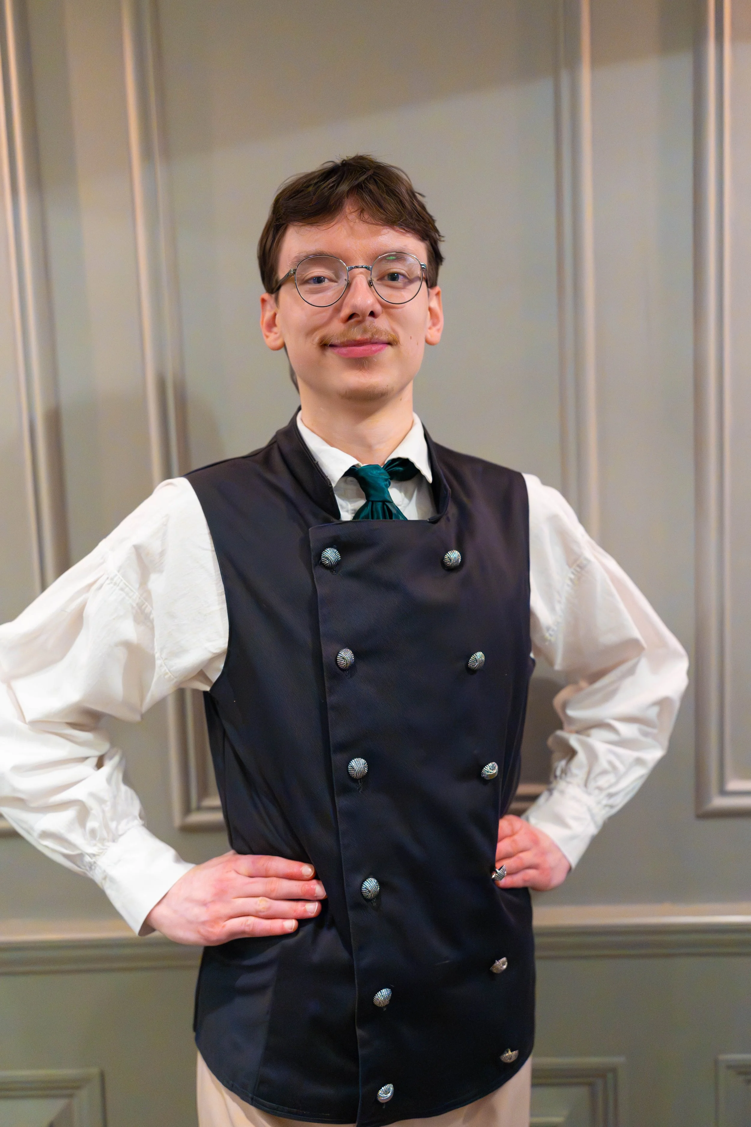 A person with short brown hair, glasses, and a mustache wearing a black double-breasted vest over a white shirt, with hands on hips, standing in front of a decorative wall panel.