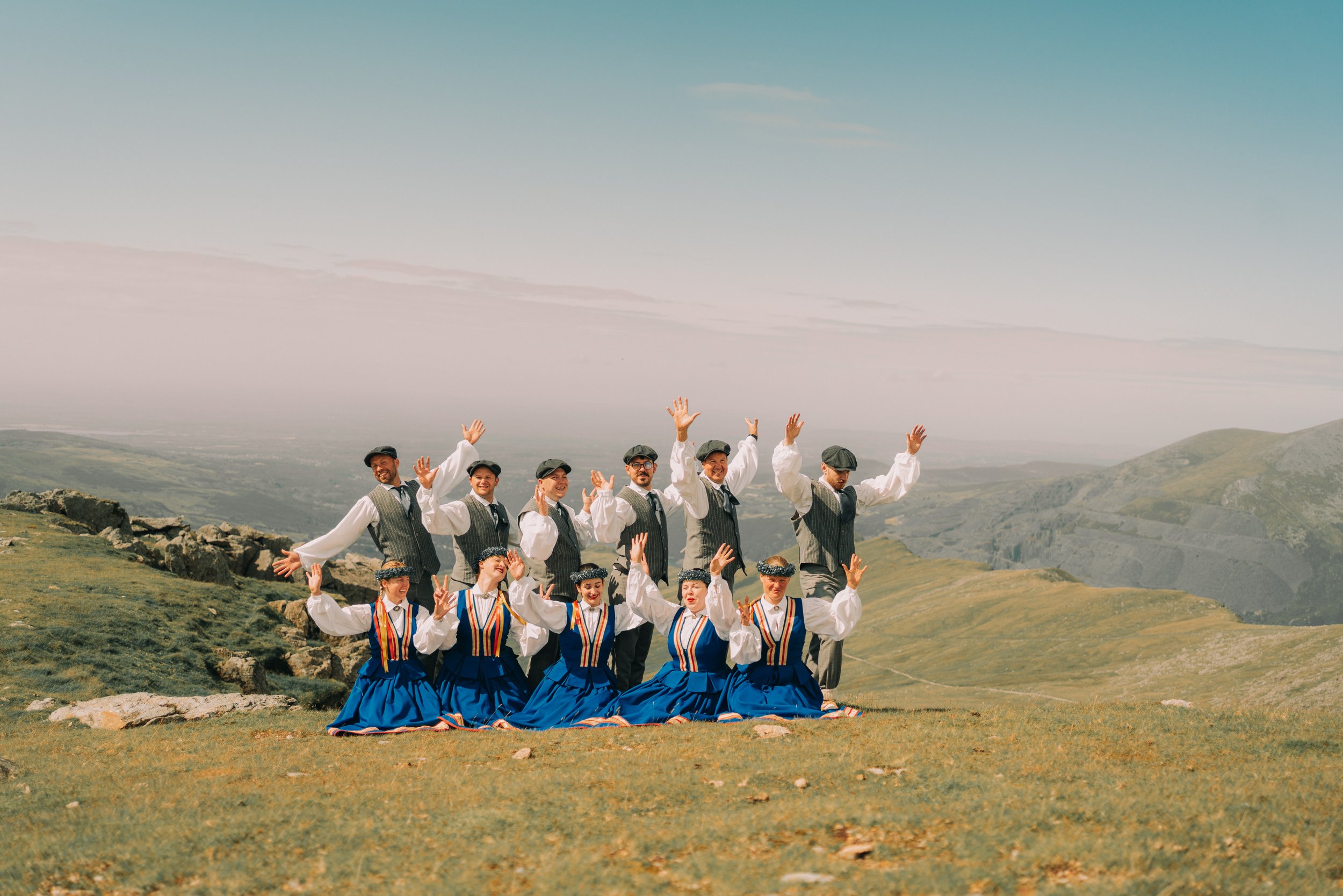 Group of people in traditional clothing outdoors, waving and smiling with mountains and a cloudy sky in the background.