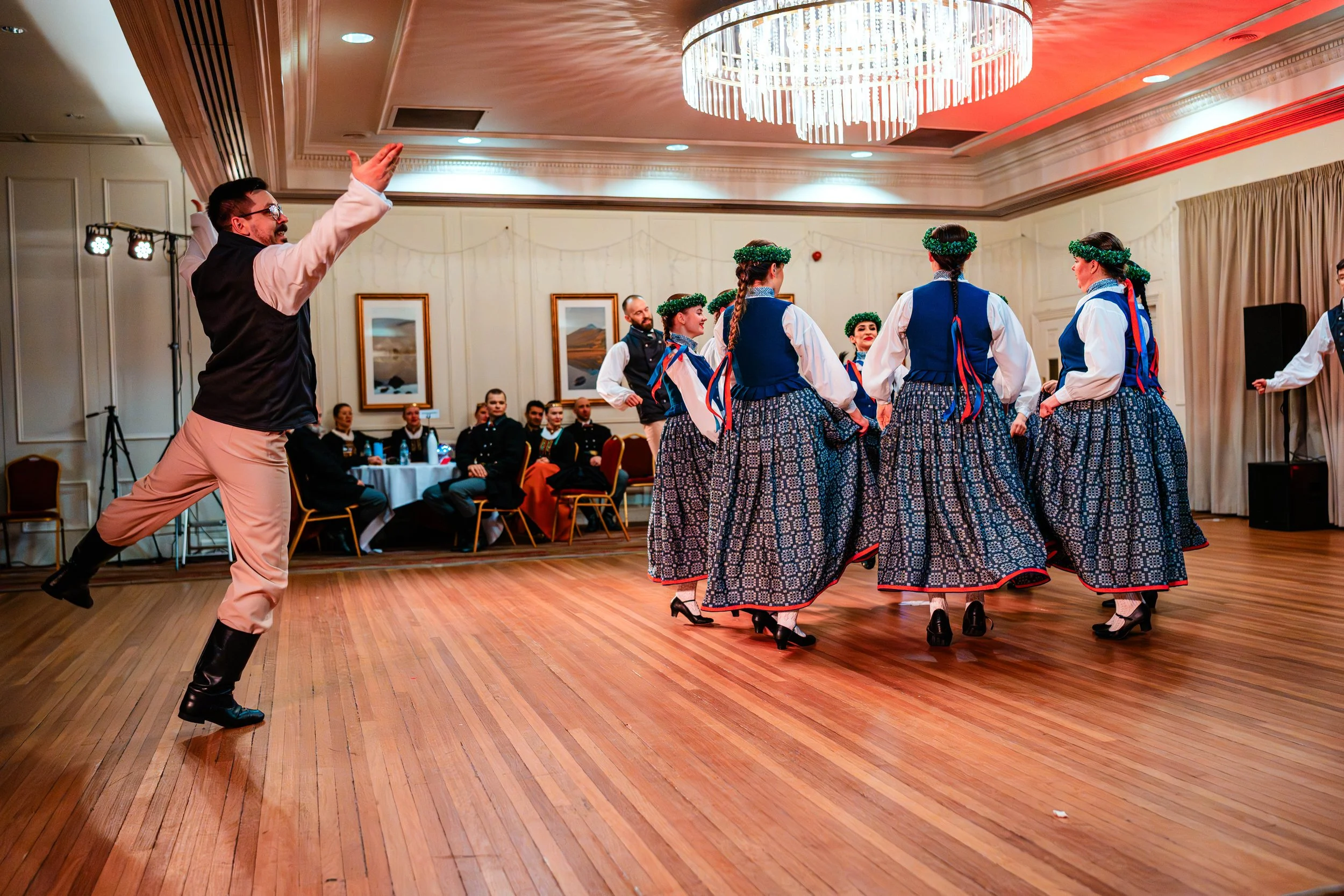 A group of performers dancing in traditional costumes in a ballroom, with an audience watching.