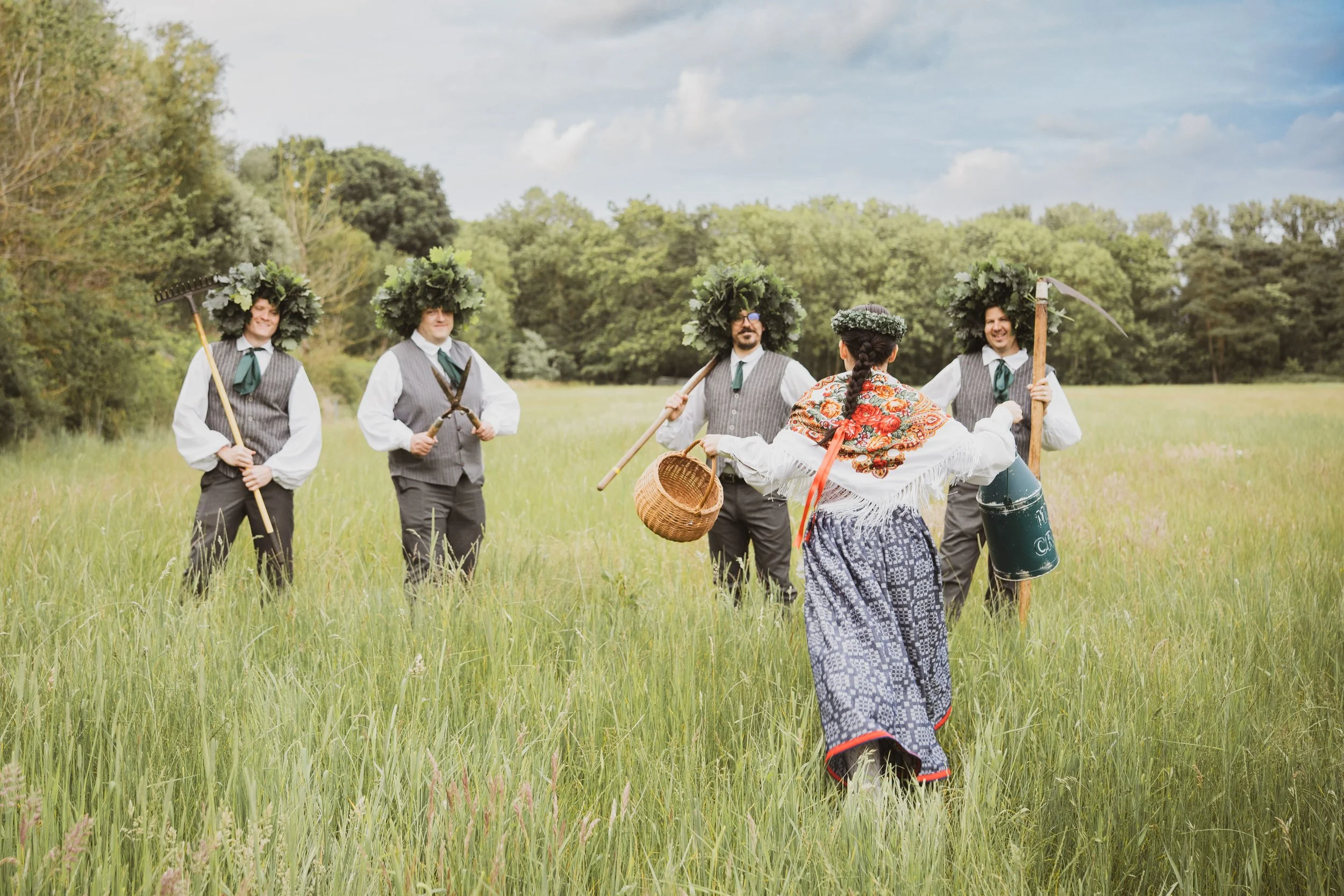 Group of five people dressed in traditional folk costumes standing in a grassy field with trees in the background, participating in a cultural or historical event.