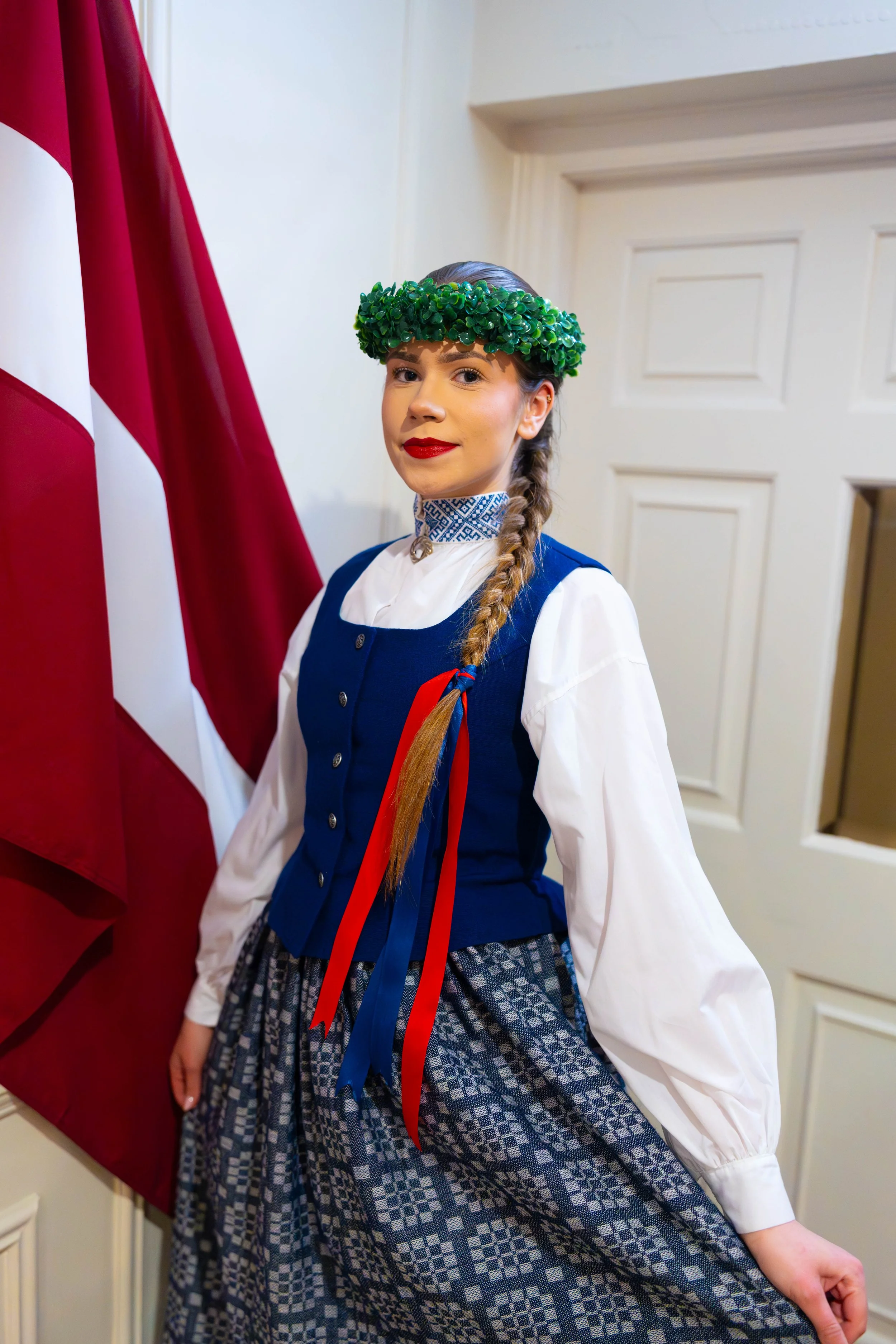 Young woman in traditional dress standing next to flag, wearing a green leafy headband and smiling.