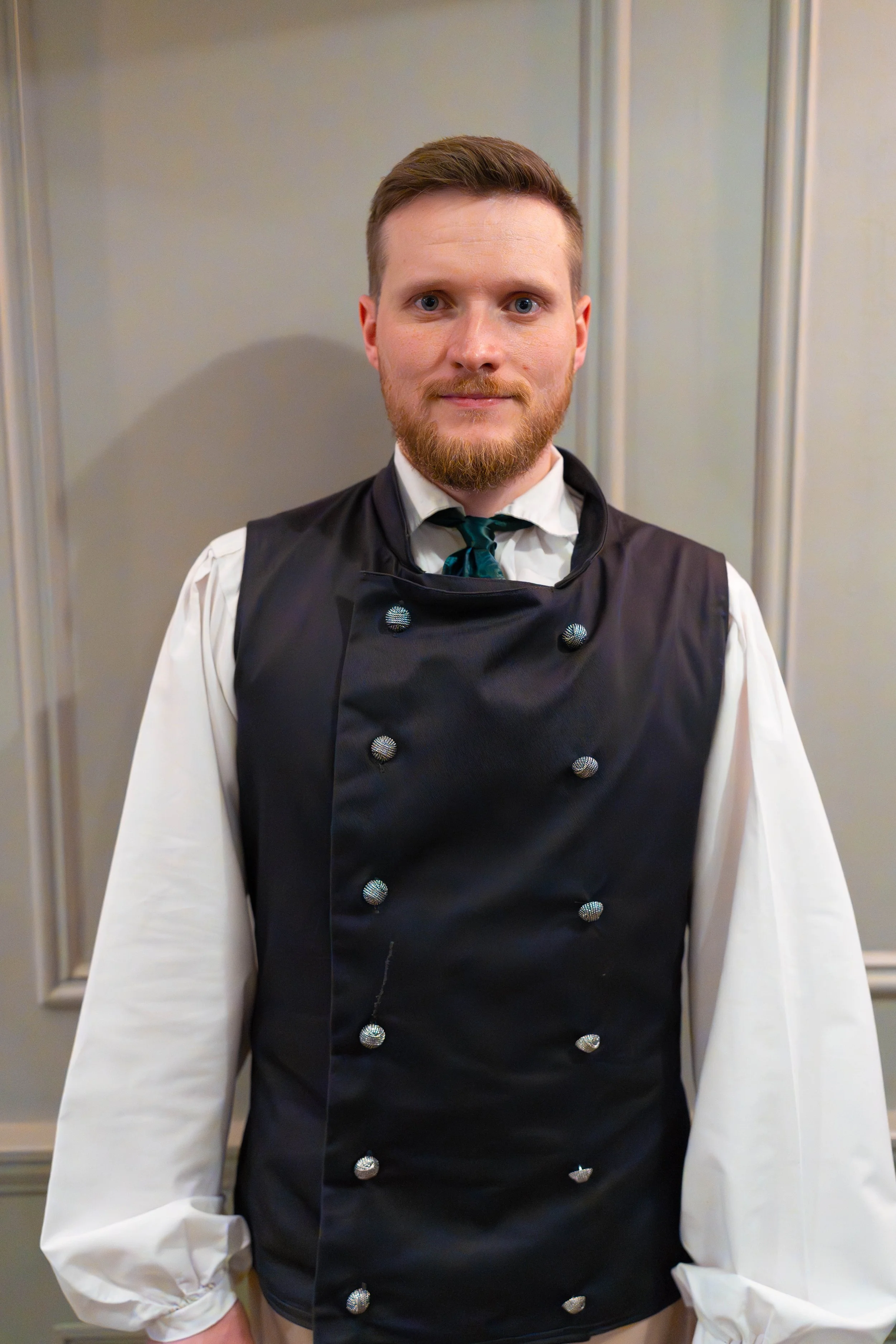 A man with light skin, a beard, and short brown hair stands in front of a plain wall. He is wearing a white shirt and a black chef's jacket with decorative buttons, along with a teal tie. His expression is neutral, and the photo appears to be taken indoors.