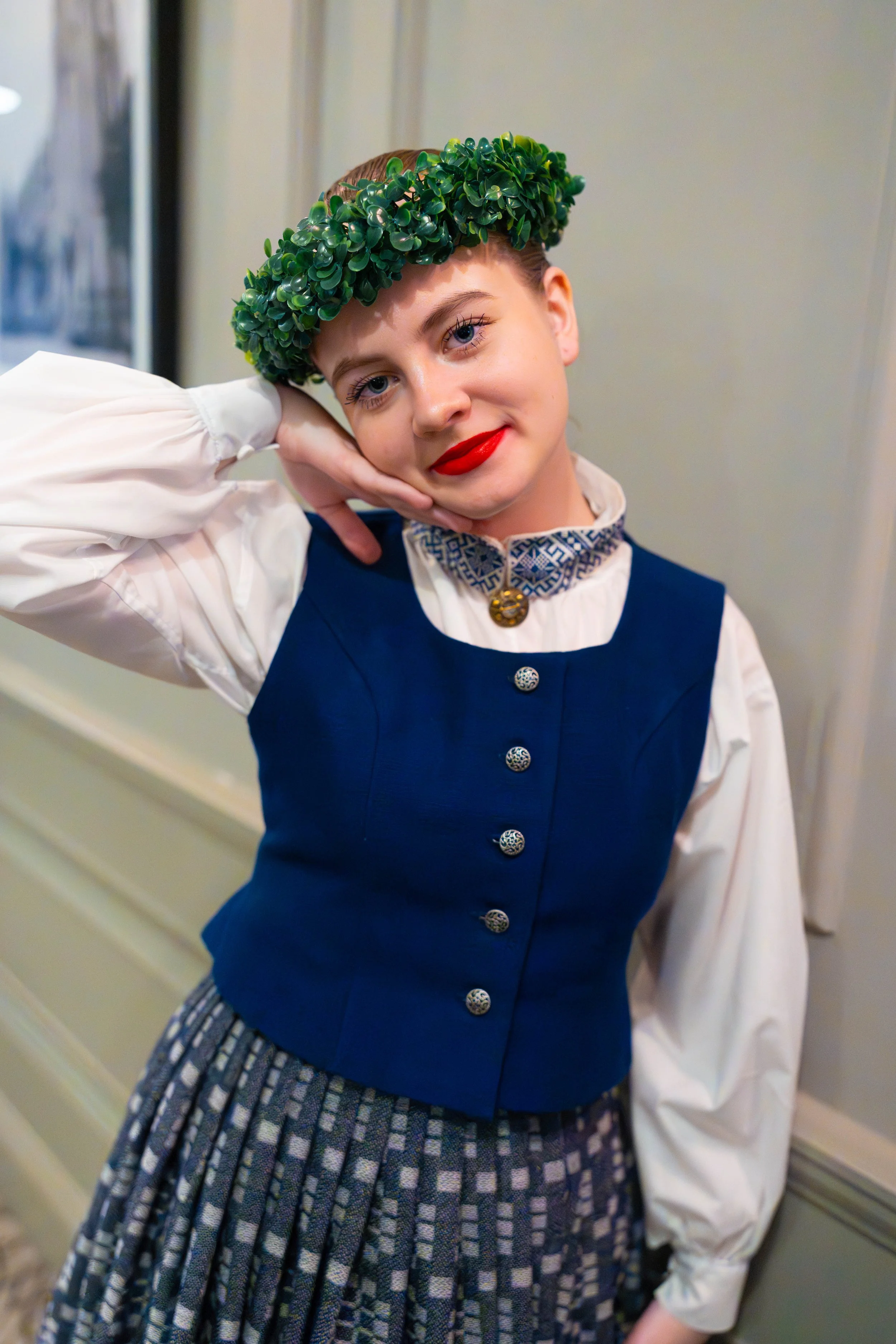 Young latvian folk dancer dressed in a traditional latvian outfit with a green headpiece, white blouse, blue vest, and patterned skirt, standing indoors and smiling.