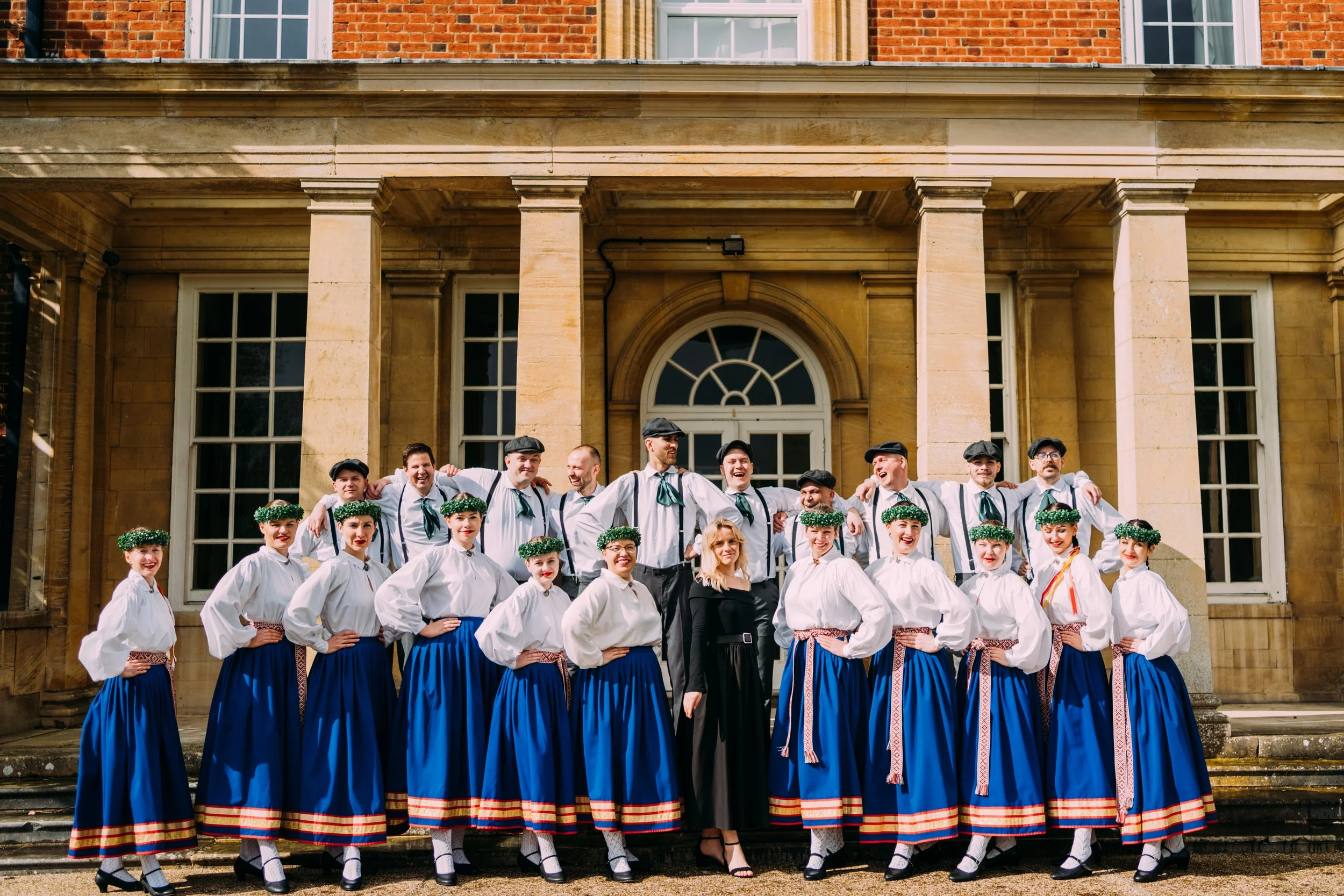 A large group of people dressed in traditional folk costumes standing on the steps of a historic building, smiling and posing for a group photo.