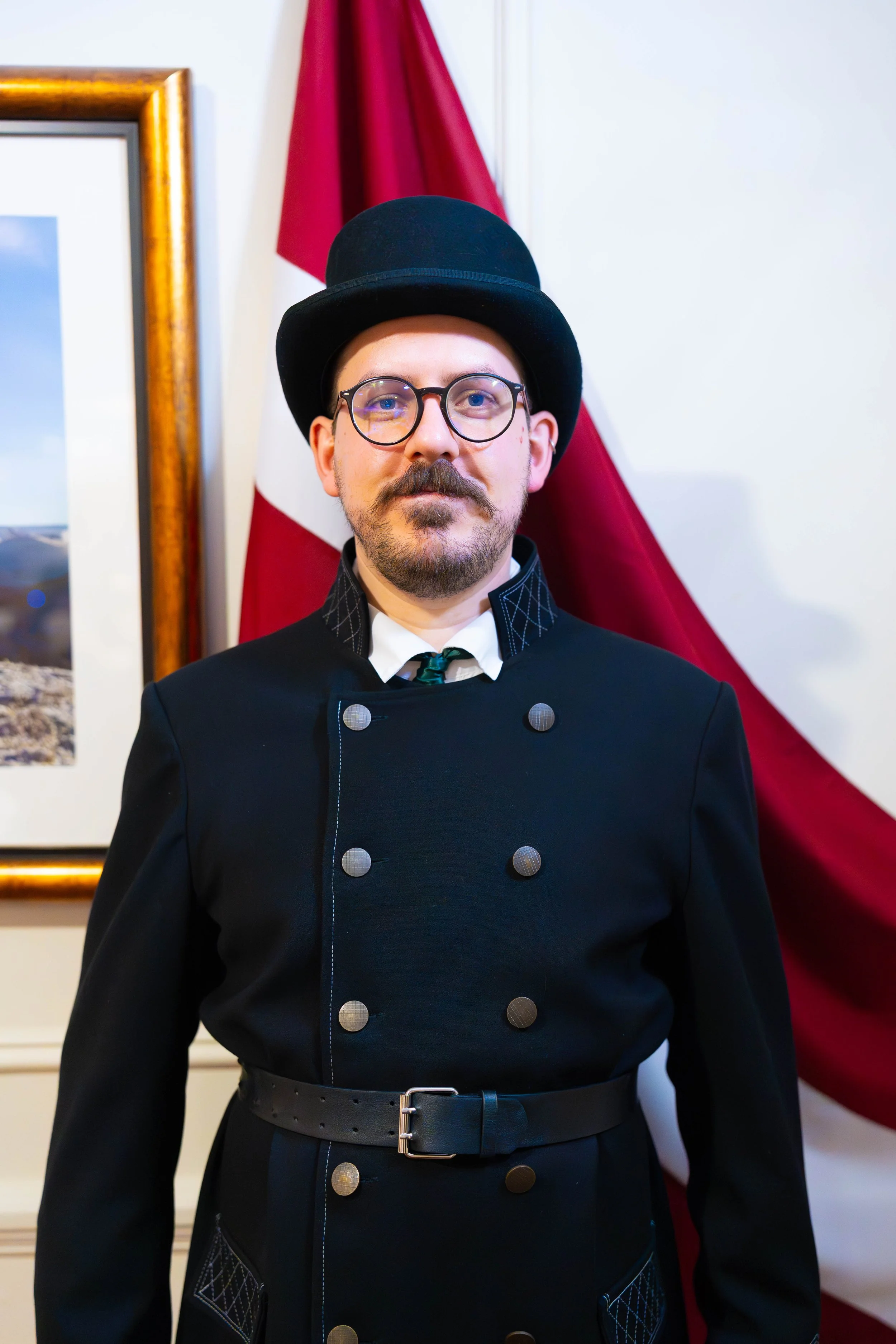 A man wearing a black chef's coat with a diamond pattern on the pockets, a wide black belt, round glasses, a mustache, and a goatee. He has a black top hat and is standing in front of an American flag, with a picture frame partially visible on the left.