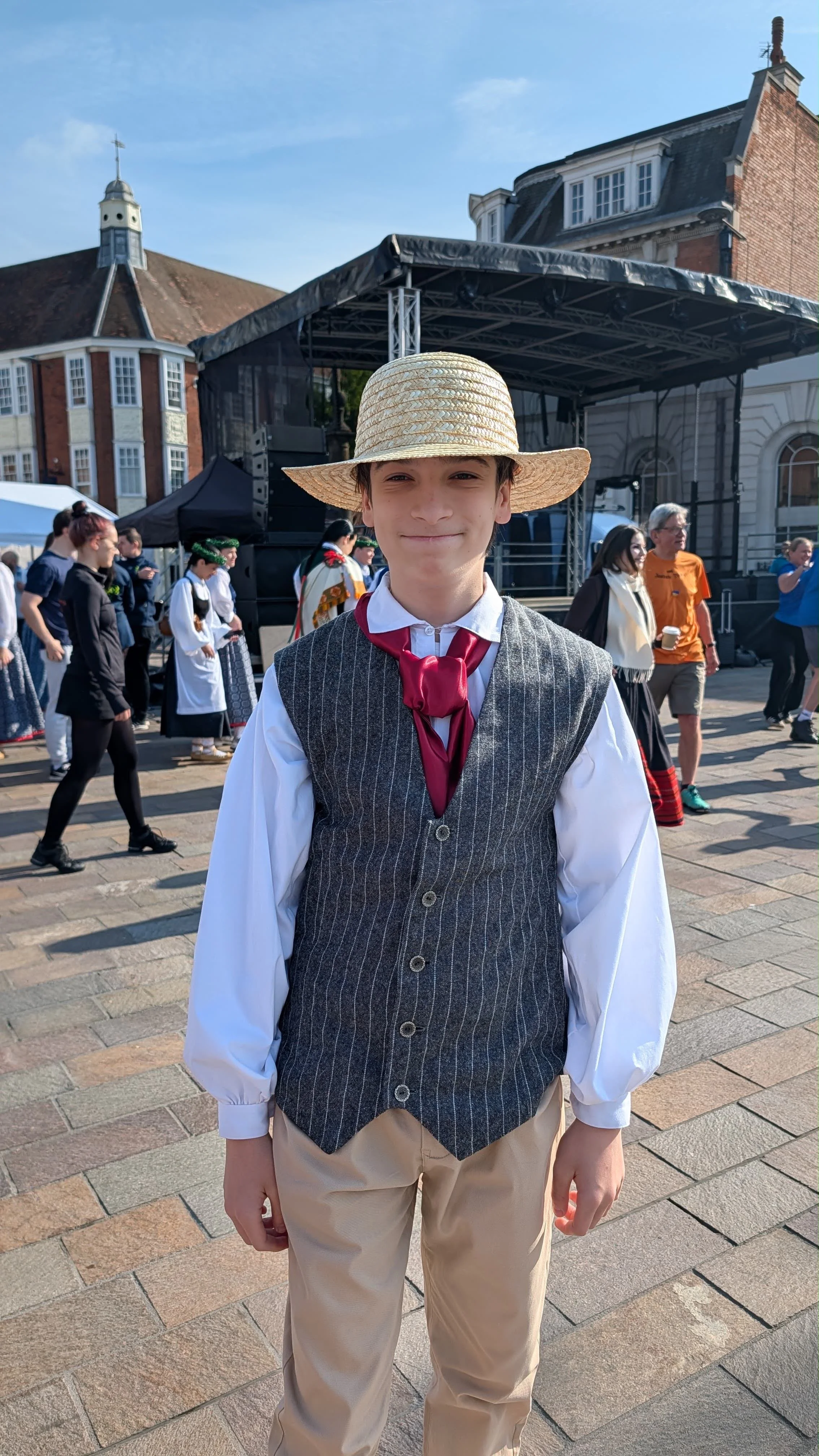 A young boy dressed in vintage-style clothing, including a straw hat, white shirt, maroon necktie, gray vest, and beige pants, standing outdoors at a cultural event or festival. There is a stage and other people dressed in traditional attire in the background, with historic buildings and a clear blue sky.