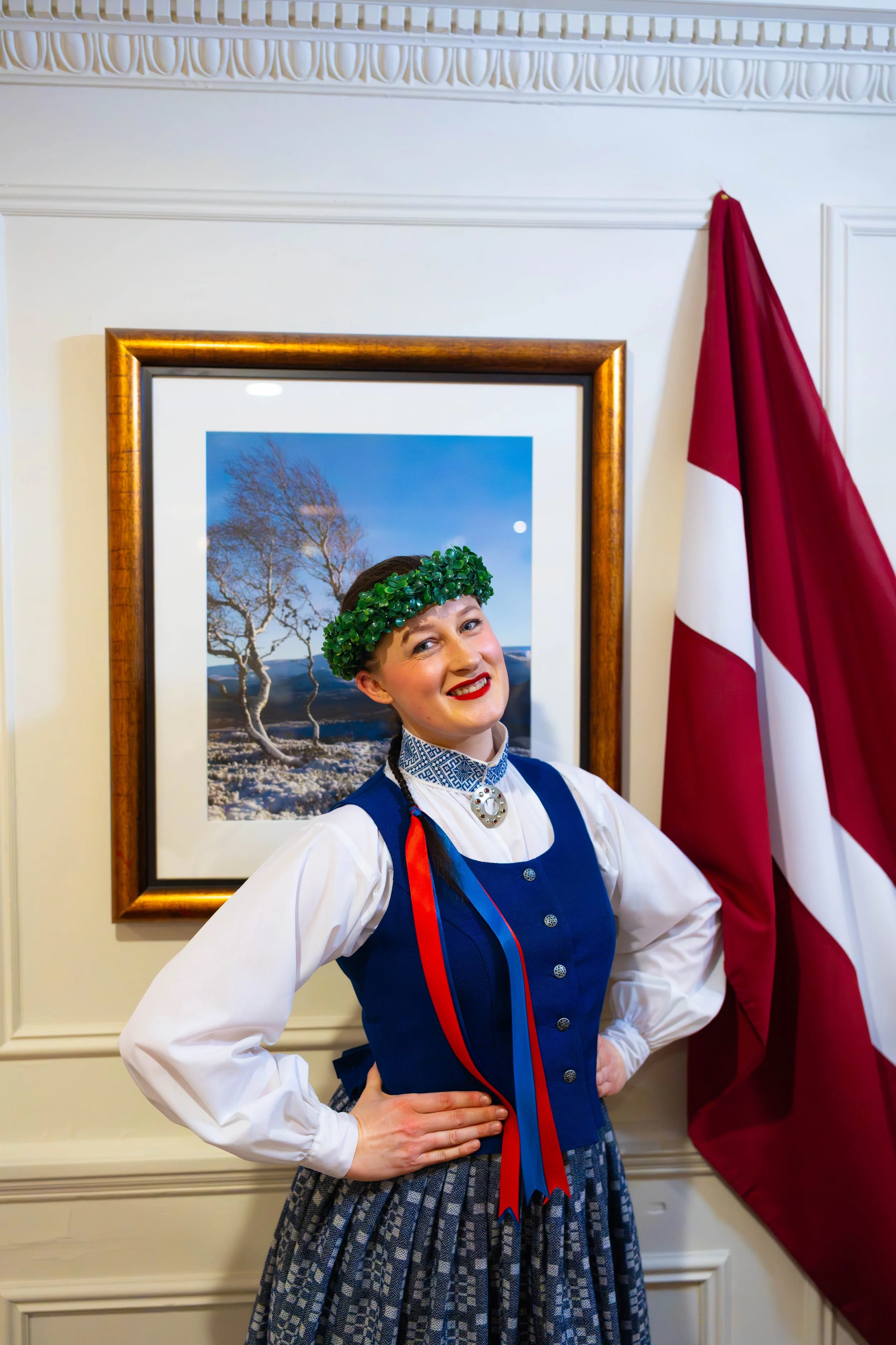 A woman dressed in traditional Norwegian clothing, called a bunad, standing in front of artwork showing snowy trees and a bright blue sky. She is wearing a green leafy crown, and has red lipstick, with her hand on her hip, smiling at the camera. A Norwegian flag is hanging on her right side.