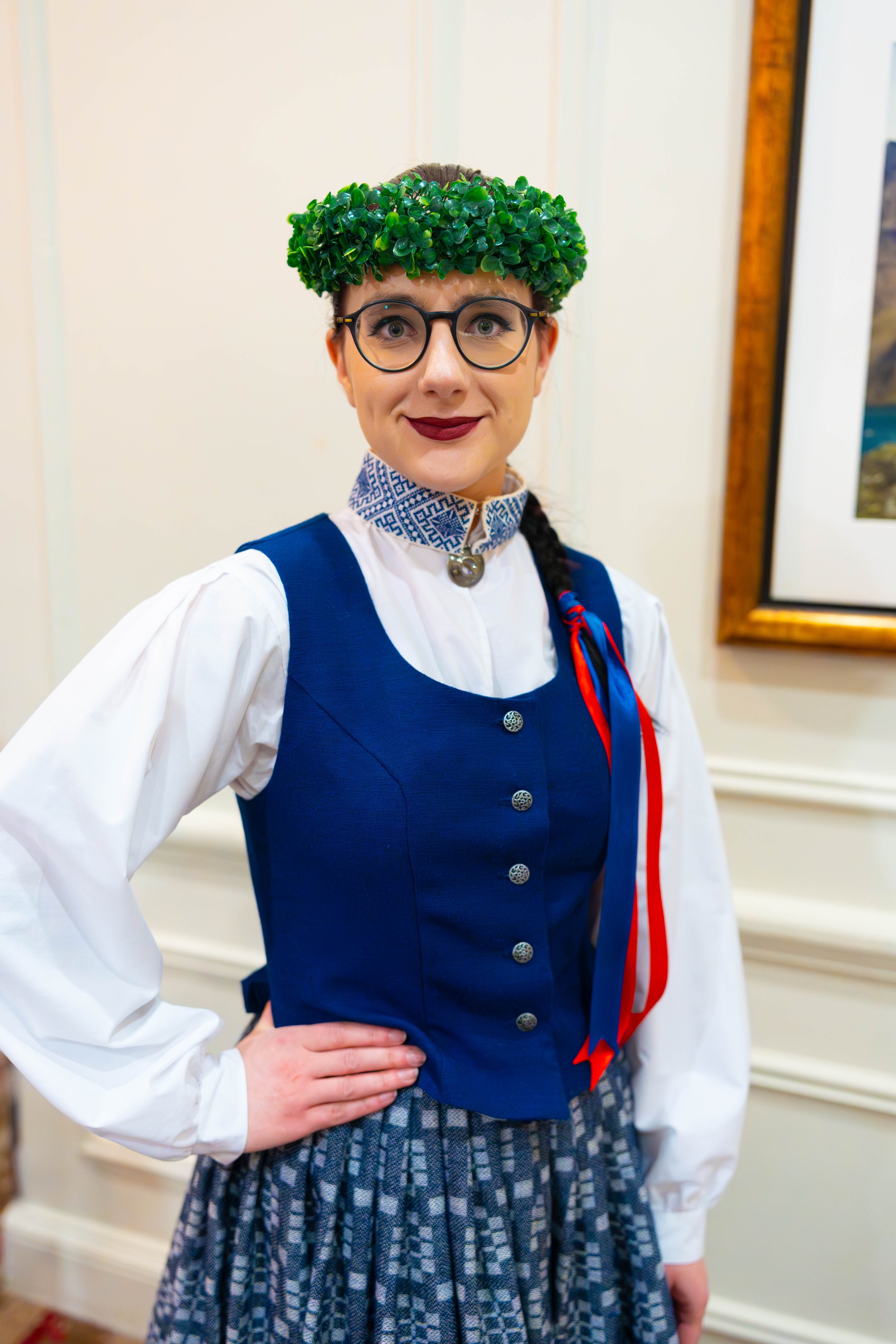 A woman dressed in traditional Scandinavian attire, wearing a blue vest with silver buttons, a white blouse with puffed sleeves, a patterned skirt, and a green floral headdress with glasses and red ribbons.