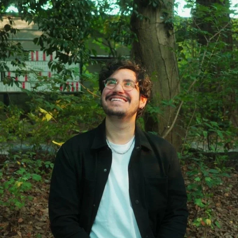 A man with glasses and a beard smiling outdoors among trees and green foliage.