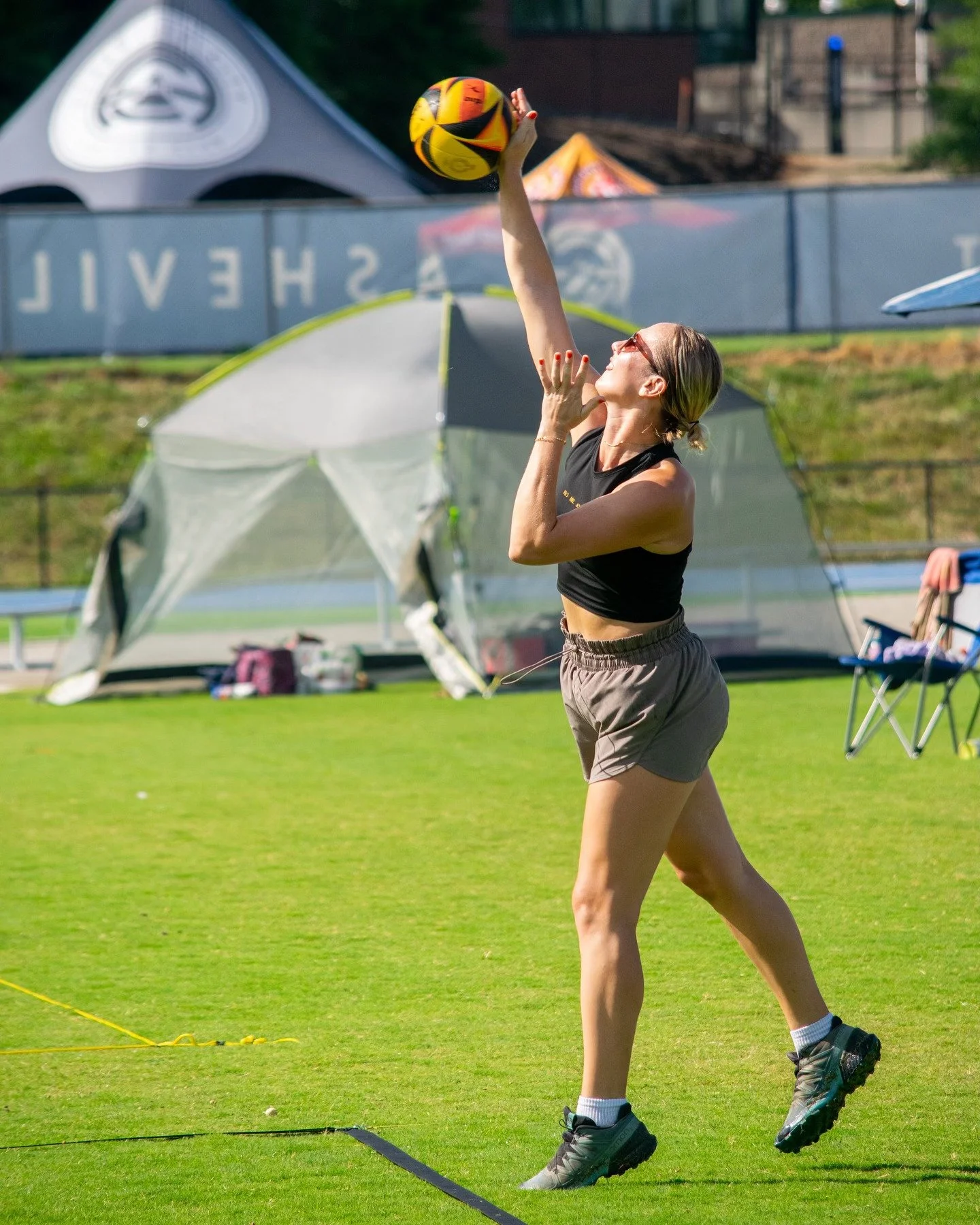 A person playing volleyball on a grassy field reaches up to hit a volleyball. They are wearing athletic clothing and sunglasses. A tent and chairs are in the background.