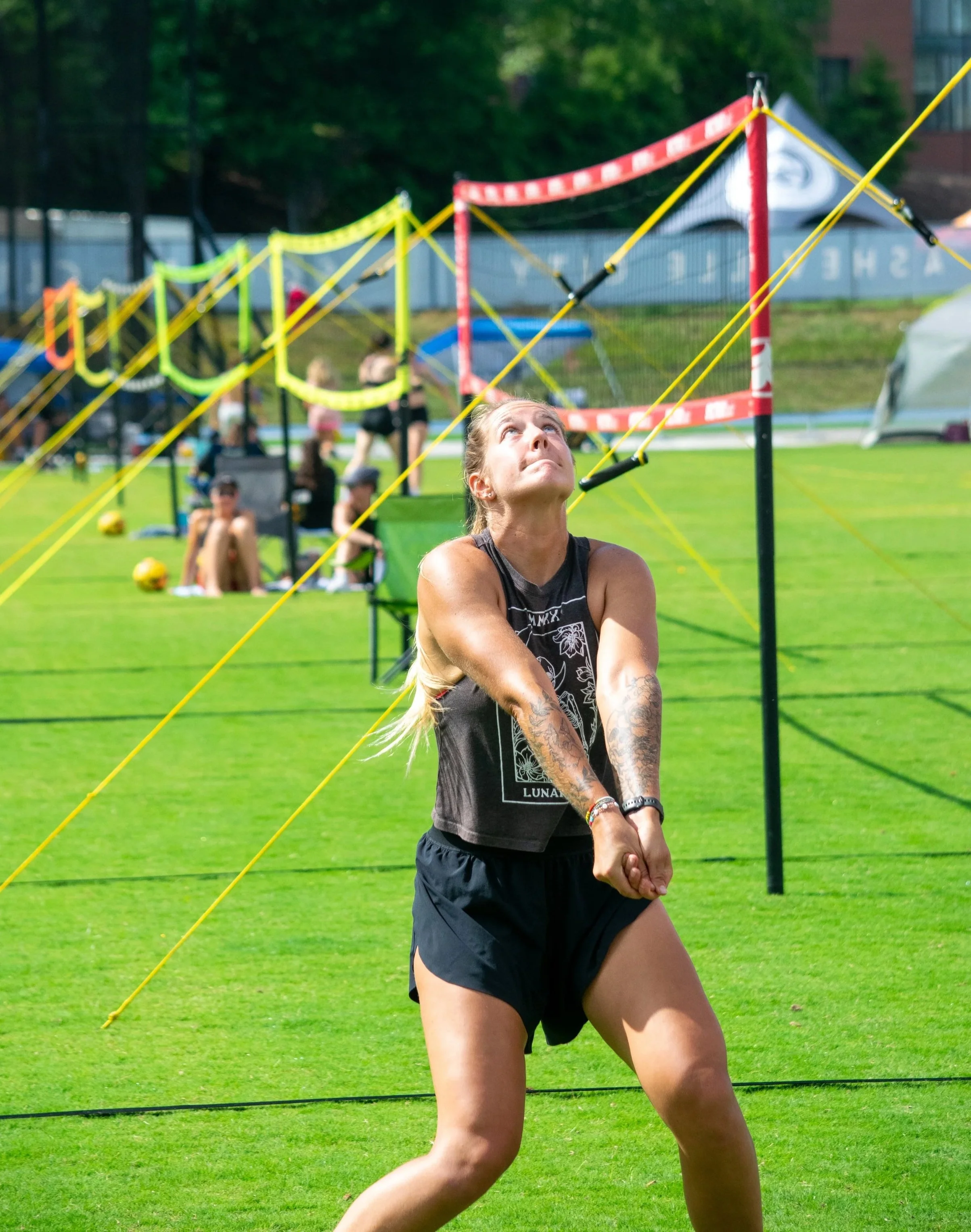 A woman with tattoos in a black tank top and shorts participating in a tug-of-war game on a grassy field during daytime, with a group of people and sports equipment in the background.