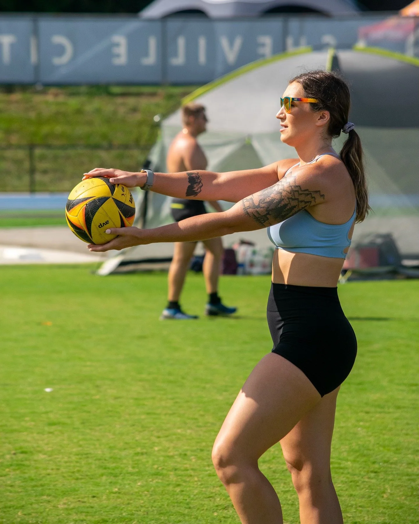 A person with a tattoo on their arm stands on a grassy field, holding a volleyball while preparing to serve. They are wearing sunglasses, a light blue top, and black shorts. In the background, another individual is visible near a tent.