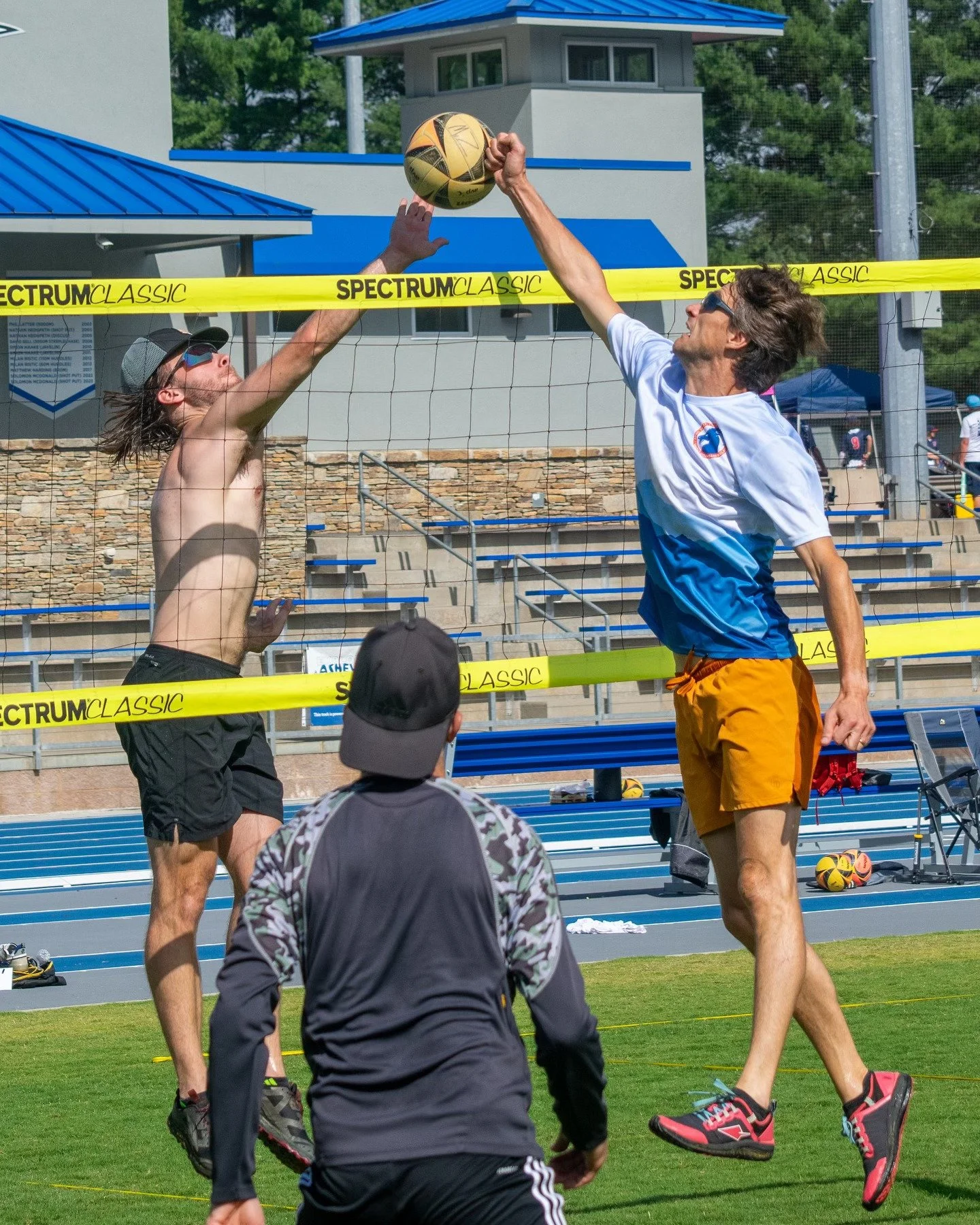 Two men playing volleyball outdoors at the beach volleyball court. One man, shirtless and with long hair, is reaching up to block the ball, while the other man in glasses, a white and blue shirt, and orange shorts is jumping to hit the ball over the net. A person in a cap and a dark jacket is watching from below.