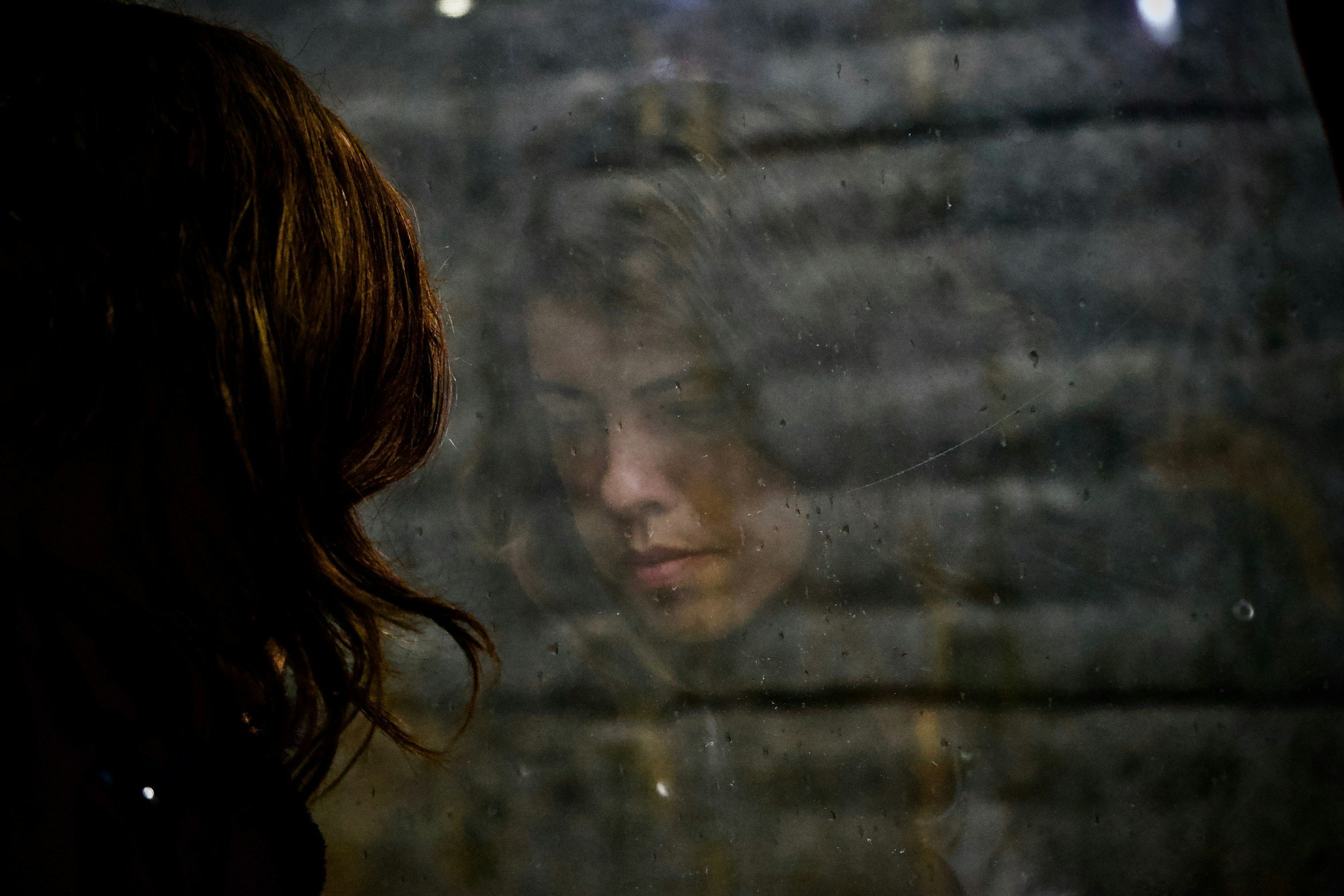 A woman with brown hair looking through a foggy or dirty window, her face reflected on the glass.