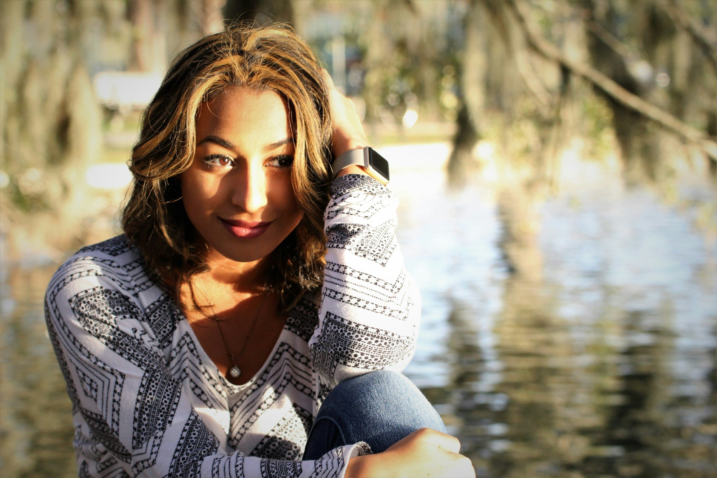 Young woman sitting outdoors near water, wearing a patterned long-sleeve shirt, with a smartwatch on her wrist, and looking at the camera.