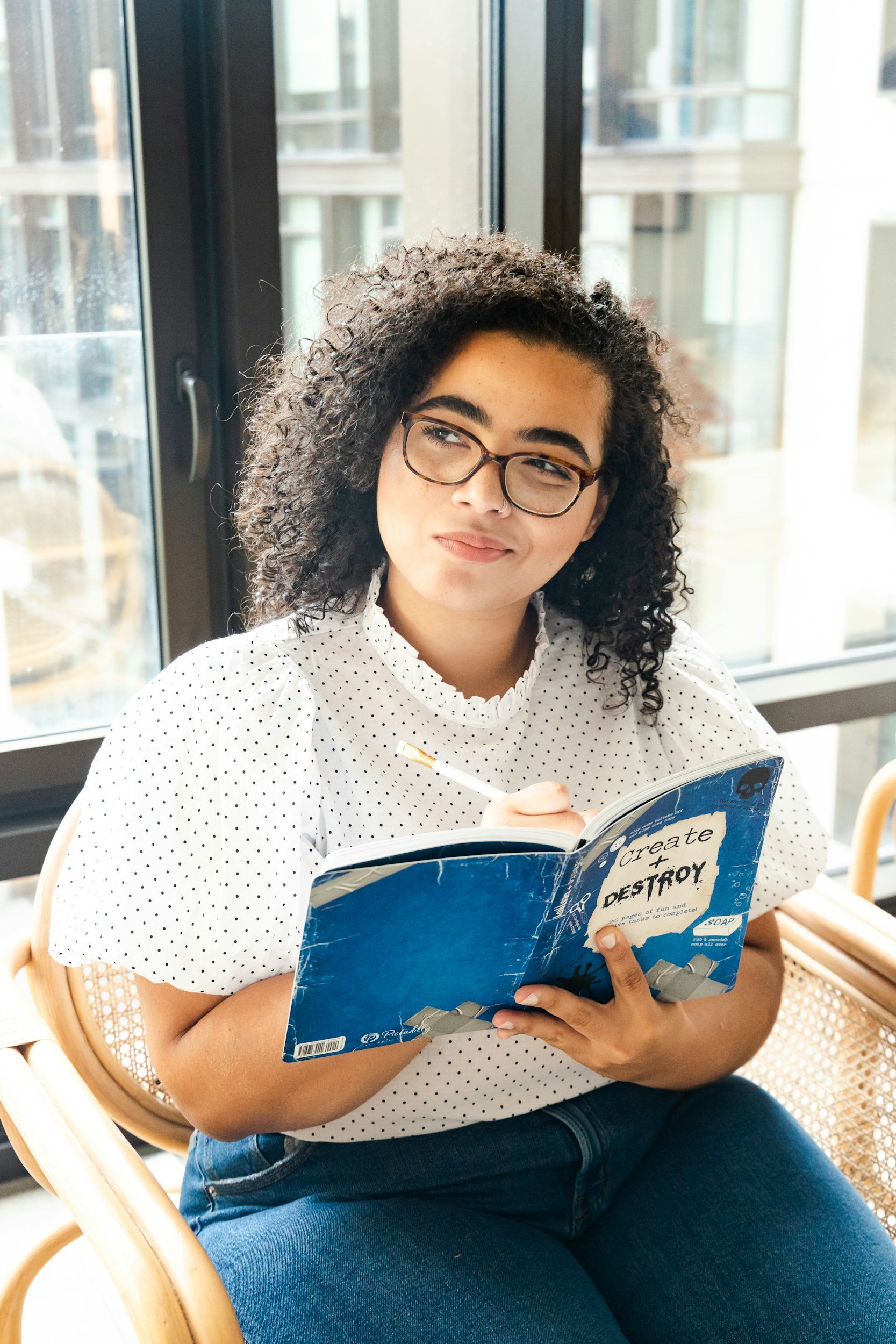 A woman wearing glasses and a white polka dot shirt sitting on a chair, holding an open book titled 'Create and Destroy' and a pencil, near large windows with a cityscape view in the background.