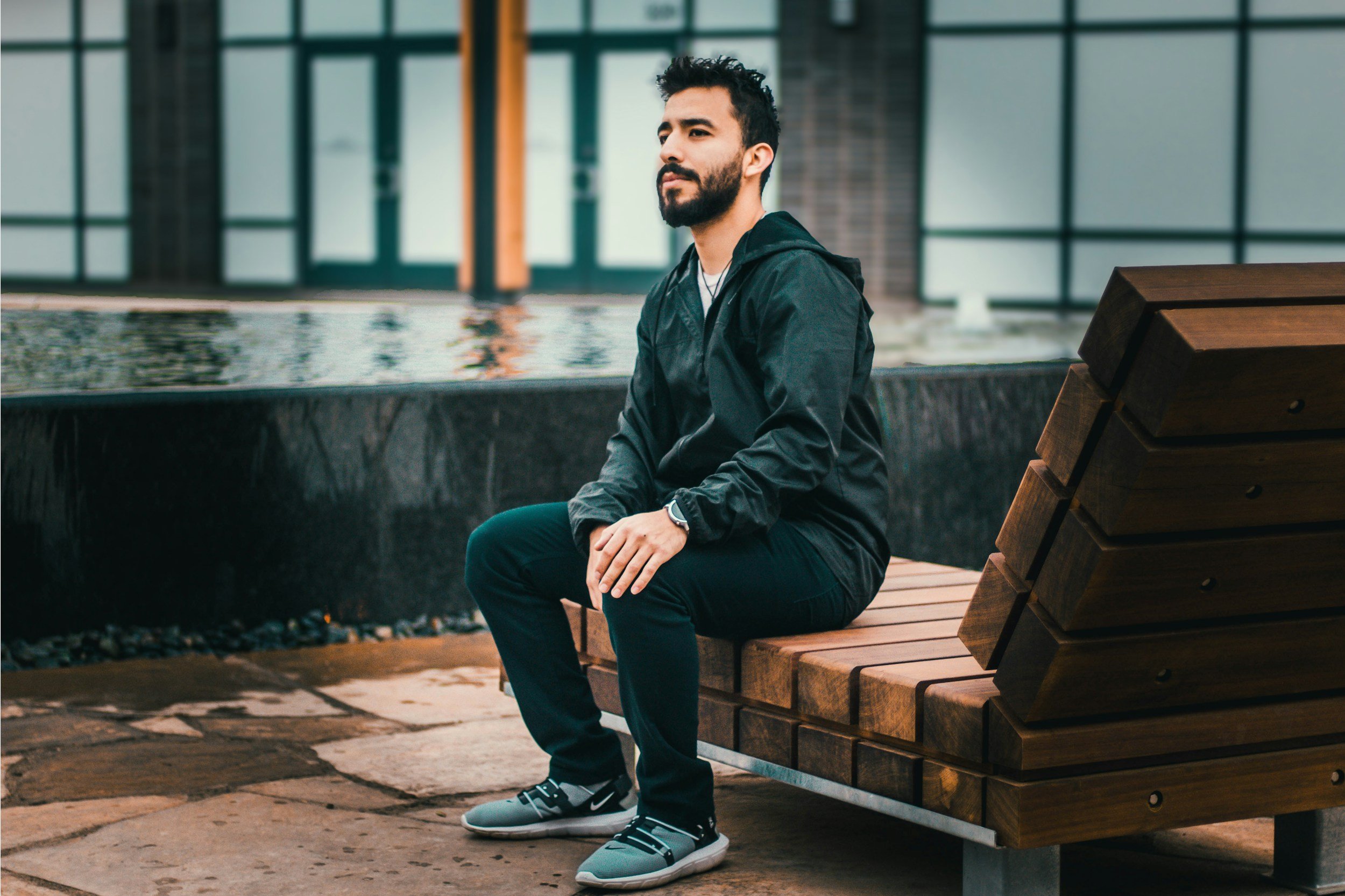 A man with a beard and dark hair sitting on a wooden park bench, wearing a black jacket, dark pants, and gray sneakers, with a modern building with large glass windows in the background.
