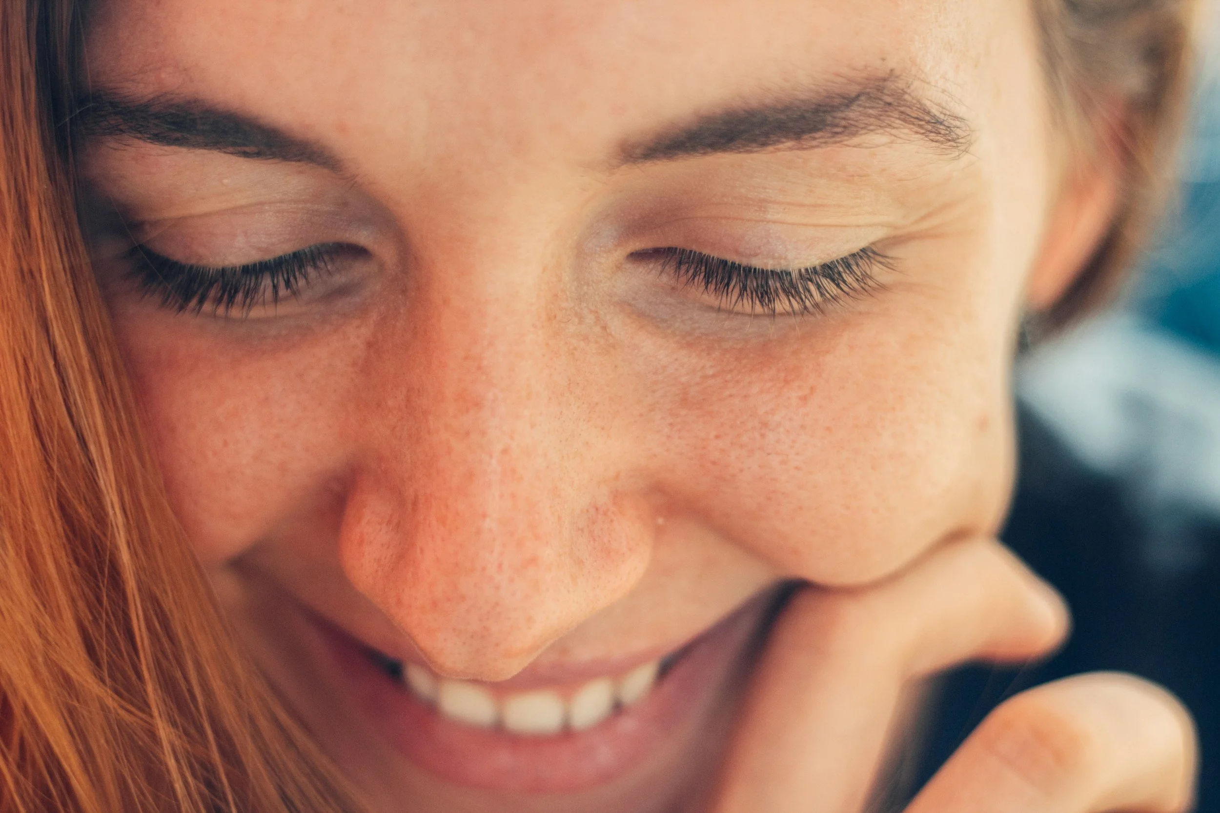 A close-up of a smiling woman with freckles, red hair, closed eyes, and natural makeup.