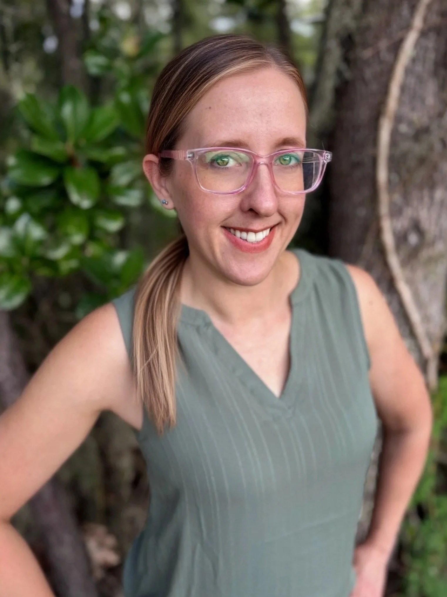 A woman with light skin, light brown hair in a low ponytail, wearing pink glasses and a sleeveless green top, standing outdoors near a tree with green foliage in the background.