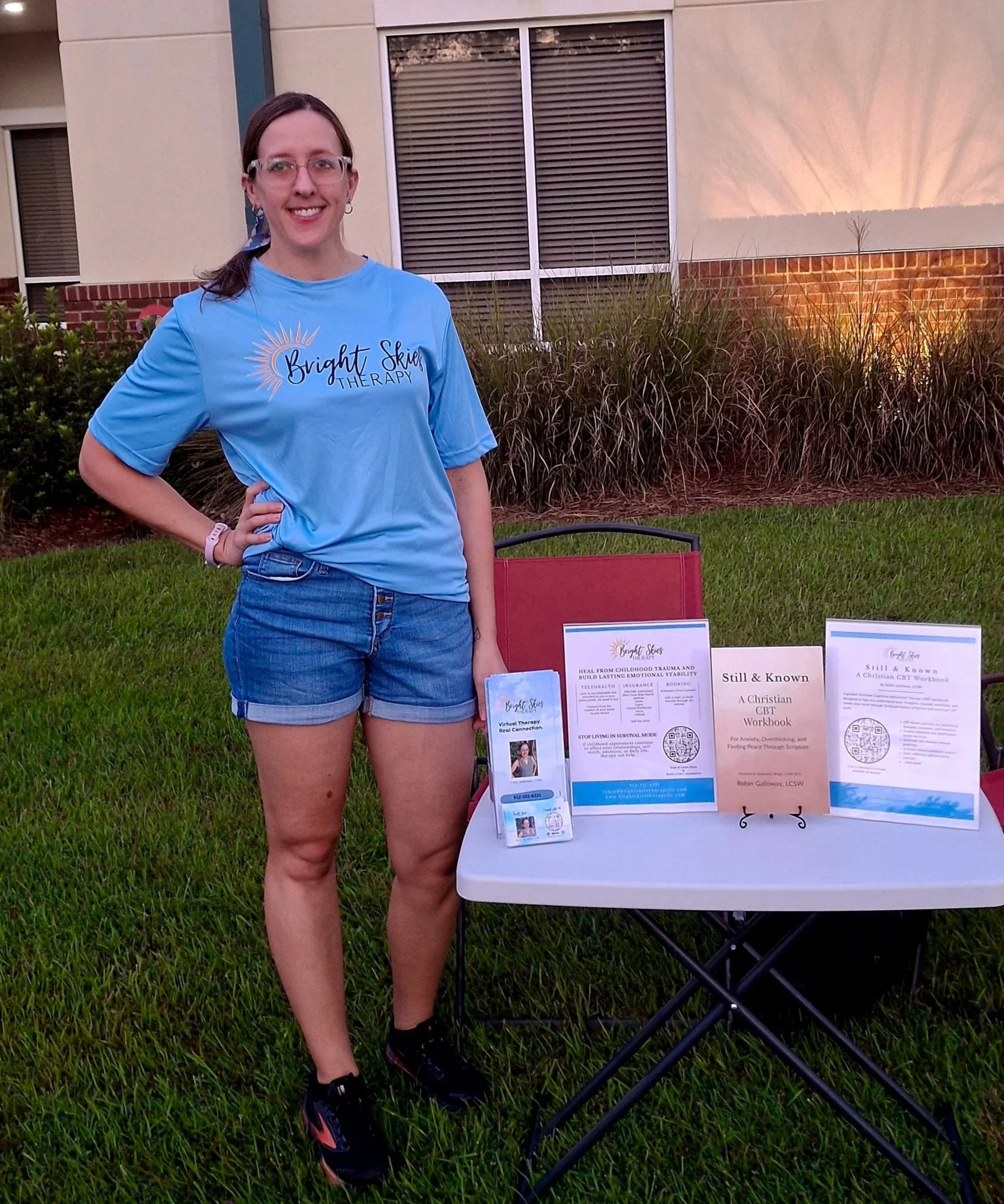 A woman wearing a bright skies therapy t-shirt standing outdoors with a display table of informational pamphlets and QR codes on mental health topics, with a building and landscaping in the background.