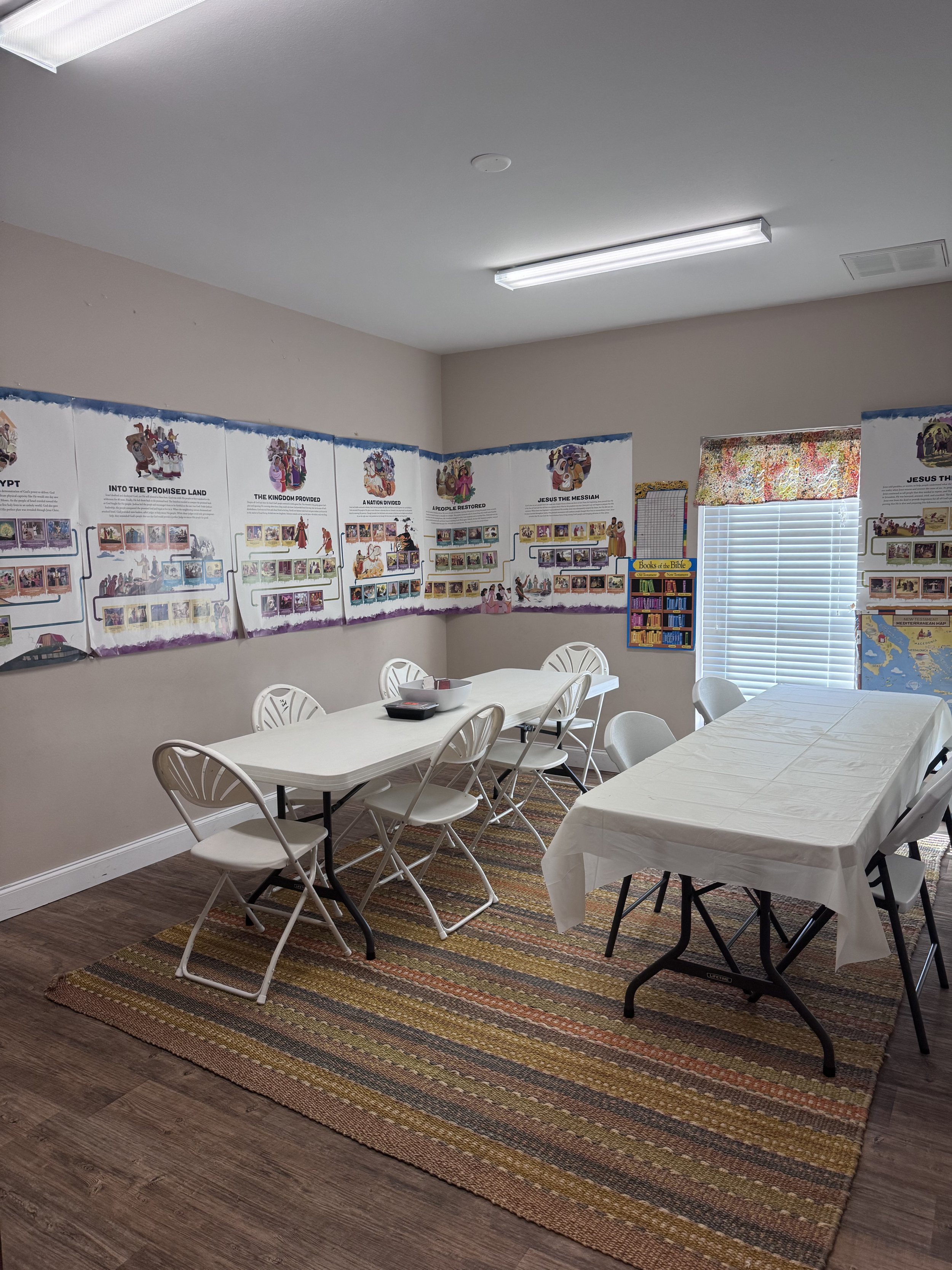 Meeting room with rectangular tables covered with white tablecloths, surrounded by white plastic chairs. The room has a colorful striped rug on a wooden floor, posters on religious themes on the walls, and a window with blinds and a floral valance. B