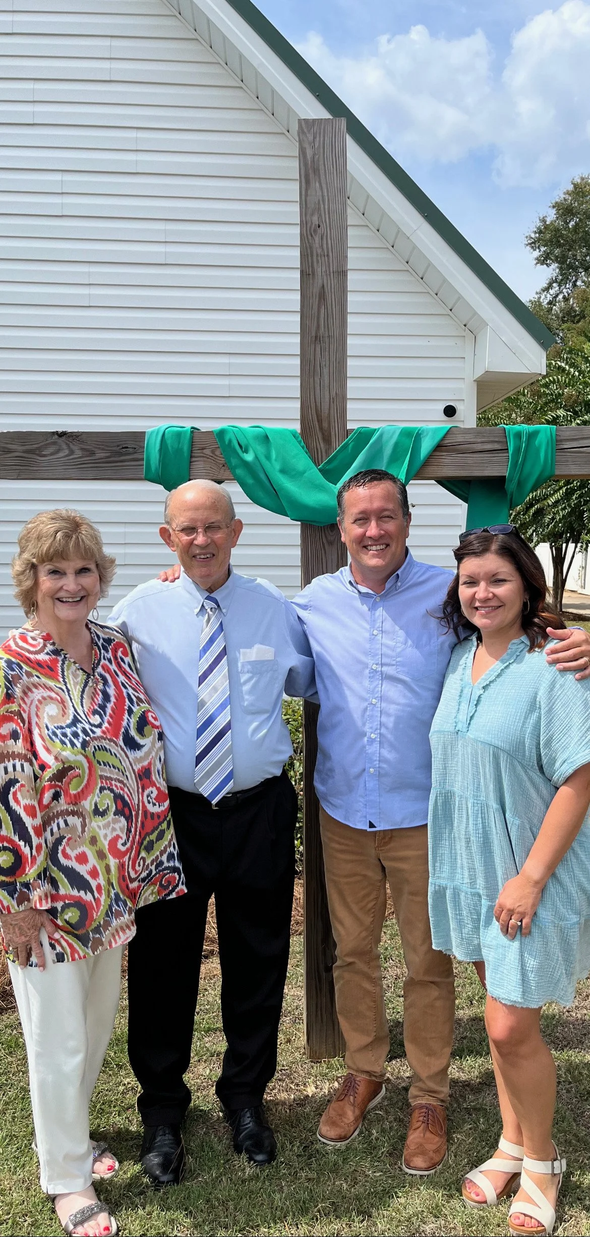 Four people standing outside in front of a wooden cross with green fabric, celebrating a religious event. The group includes two women and two men, smiling, dressed in casual and semi-formal clothing. A white building with siding is in the background