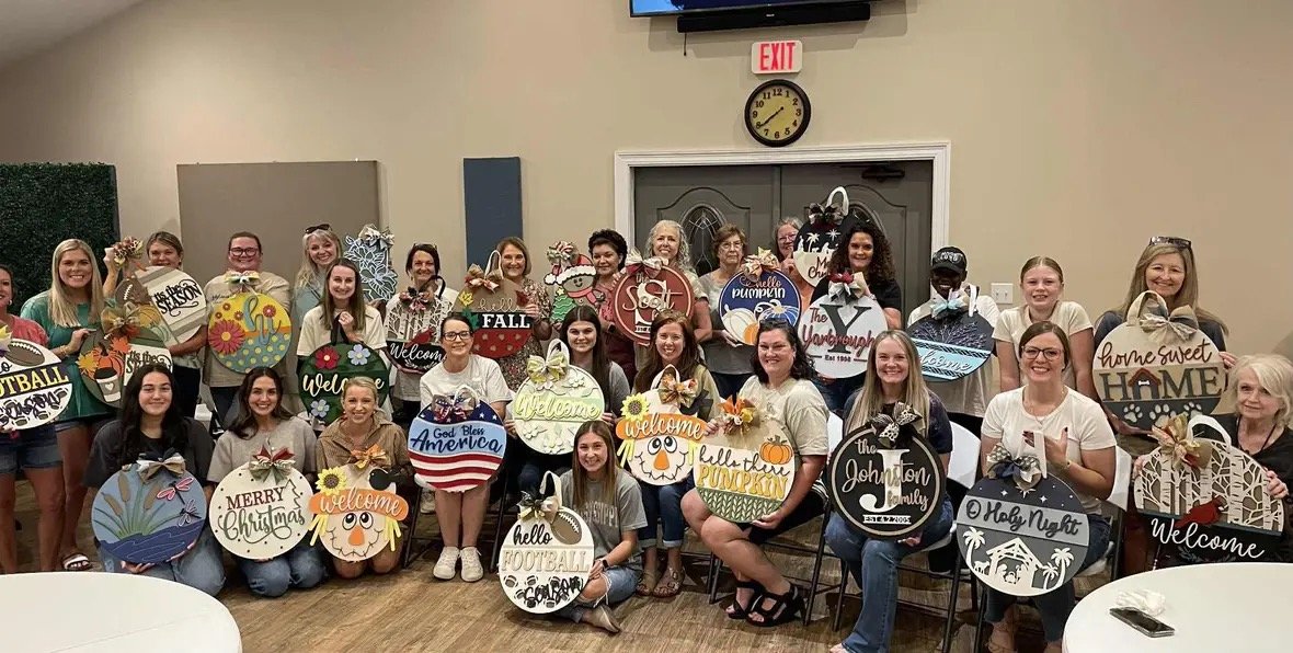 A group of women and girls holding decorated signs with festive and seasonal themes, posing indoors with a wall, clock, and exit sign in the background.